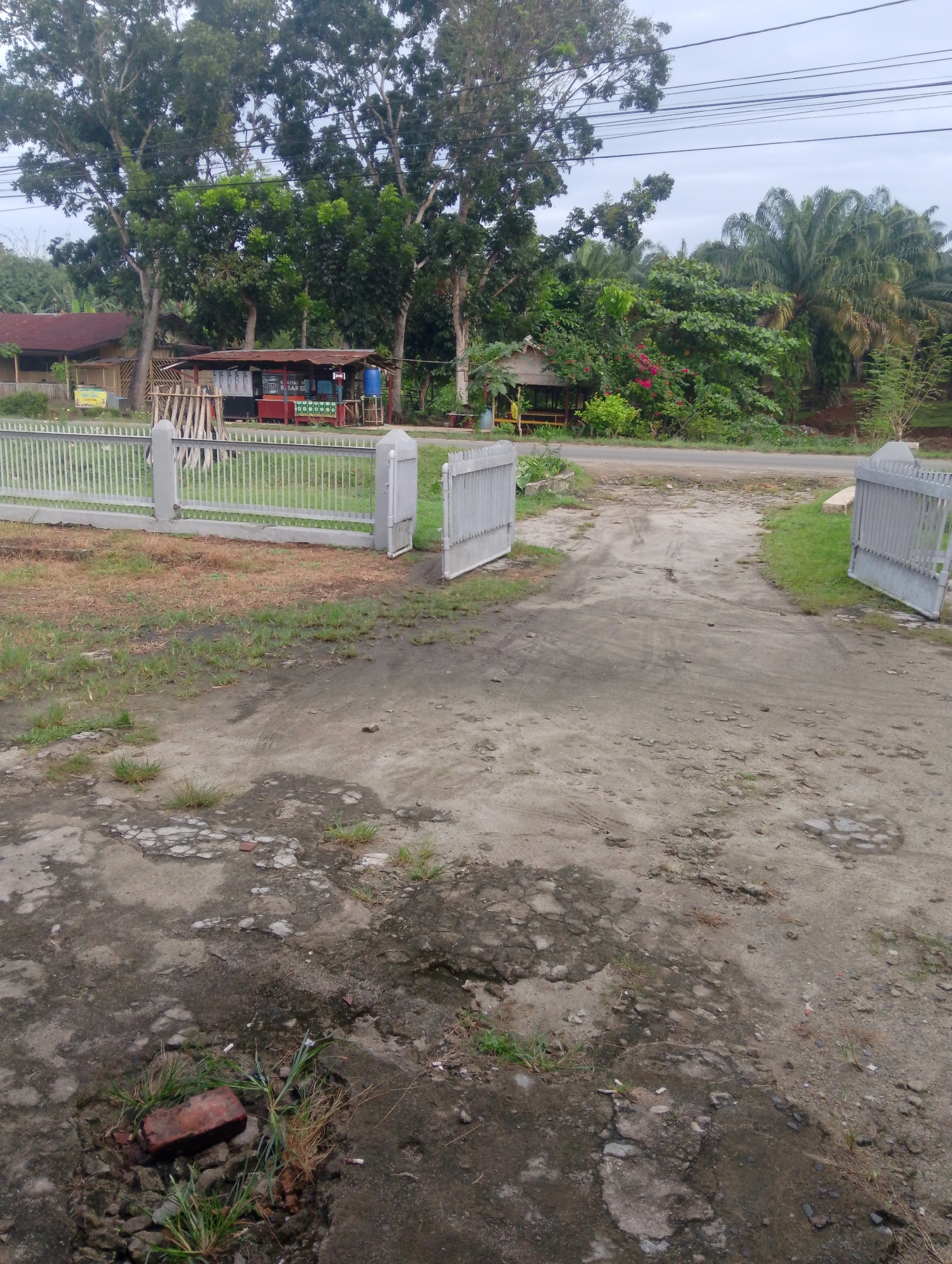 The image depicts an outdoor scene in Serbelawan, Indonesia. A grey metal fence with a white concrete base extends across the foreground, with two open gates leading to a dirt driveway. The driveway is in disrepair, with patches of exposed dirt and gravel. To the left of the driveway is a small, open-air food stall with a red counter and a corrugated metal roof. Behind the stall, a building with a brown roof is visible. To the right of the driveway, the ground is covered with patchy green grass. In the background, lush green trees and palm trees dominate the landscape, with a paved road visible in the distance. Power lines stretch across the upper portion of the image. The sky is overcast.