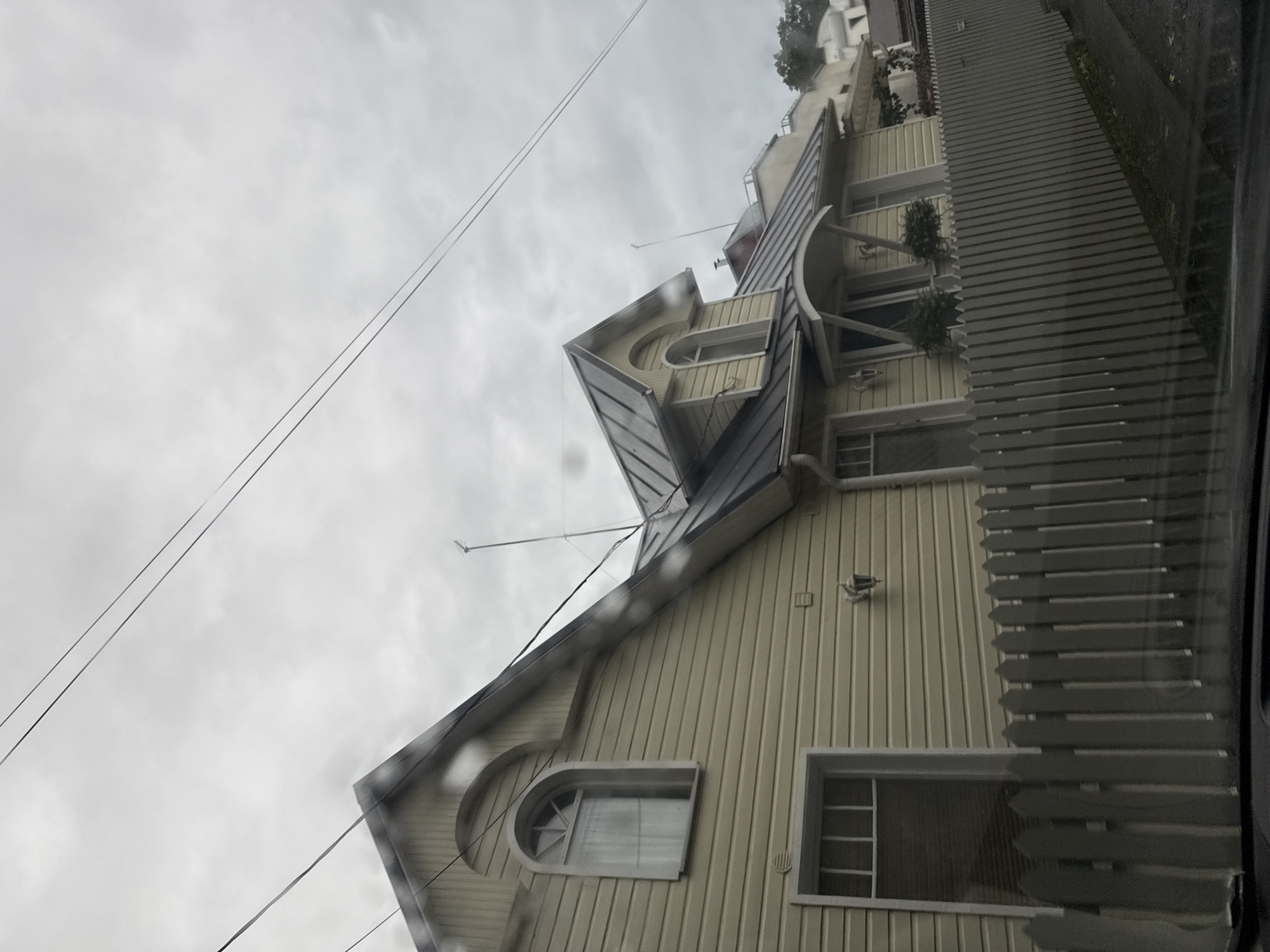 The image shows a pale yellow house with a dark gray metal roof, viewed from a low angle. The house has a picket fence in front of it. There are two windows visible on the left side of the house, one arched and the other rectangular. There are also lights mounted on the exterior of the house. The sky is overcast and gray. There are power lines visible in the upper portion of the image. The scene appears to be in a residential area. The image does not depict the city of Tallinn, Estonia.