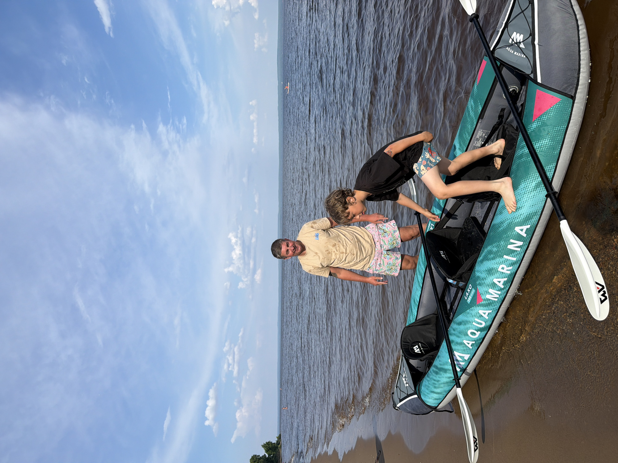 In Noviy, Russia, on a bright, sunny day with scattered clouds, two individuals, a man and a boy, are standing by an inflatable kayak partially submerged in a large body of water. The man, wearing a light-colored t-shirt and patterned shorts, stands near the rear of the kayak, facing the camera and smiling. The boy, dressed in a black t-shirt and patterned shorts, is positioned within the kayak, bending over and appearing to be preparing to embark. The kayak, teal and gray with "AQUA MARINA" branding, rests on a sandy beach. Two white paddles with black shafts lie across the kayak and in the water. The background features a wide expanse of water stretching to a distant tree line under a vast blue sky.