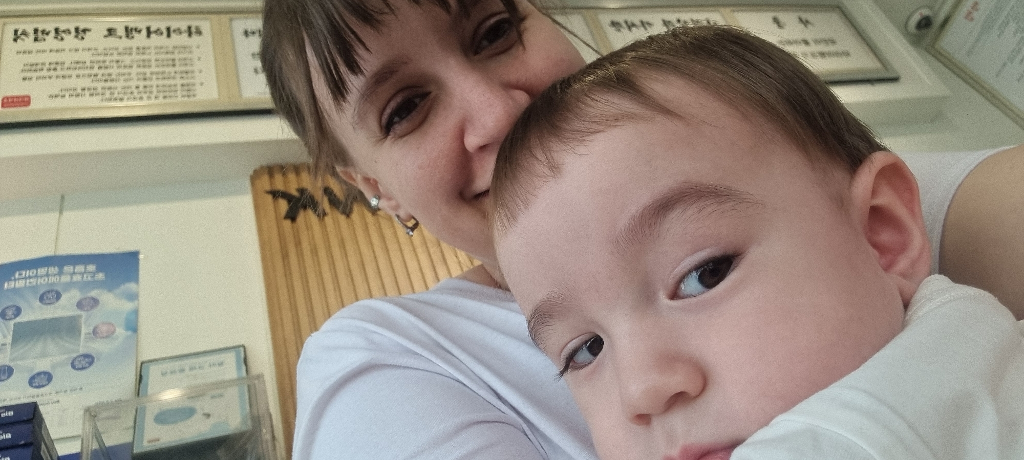 A woman is holding a child in an indoor setting in Wonju-si, South Korea. The woman has short brown hair with bangs, a light complexion, and is wearing a white t-shirt and earrings. The child has light skin, brown hair, and brown eyes, also wearing a white shirt. Behind the woman are a light-colored wall with framed informational posters in Korean, a wooden panel with a dark design, and what appears to be a display case with additional signage.