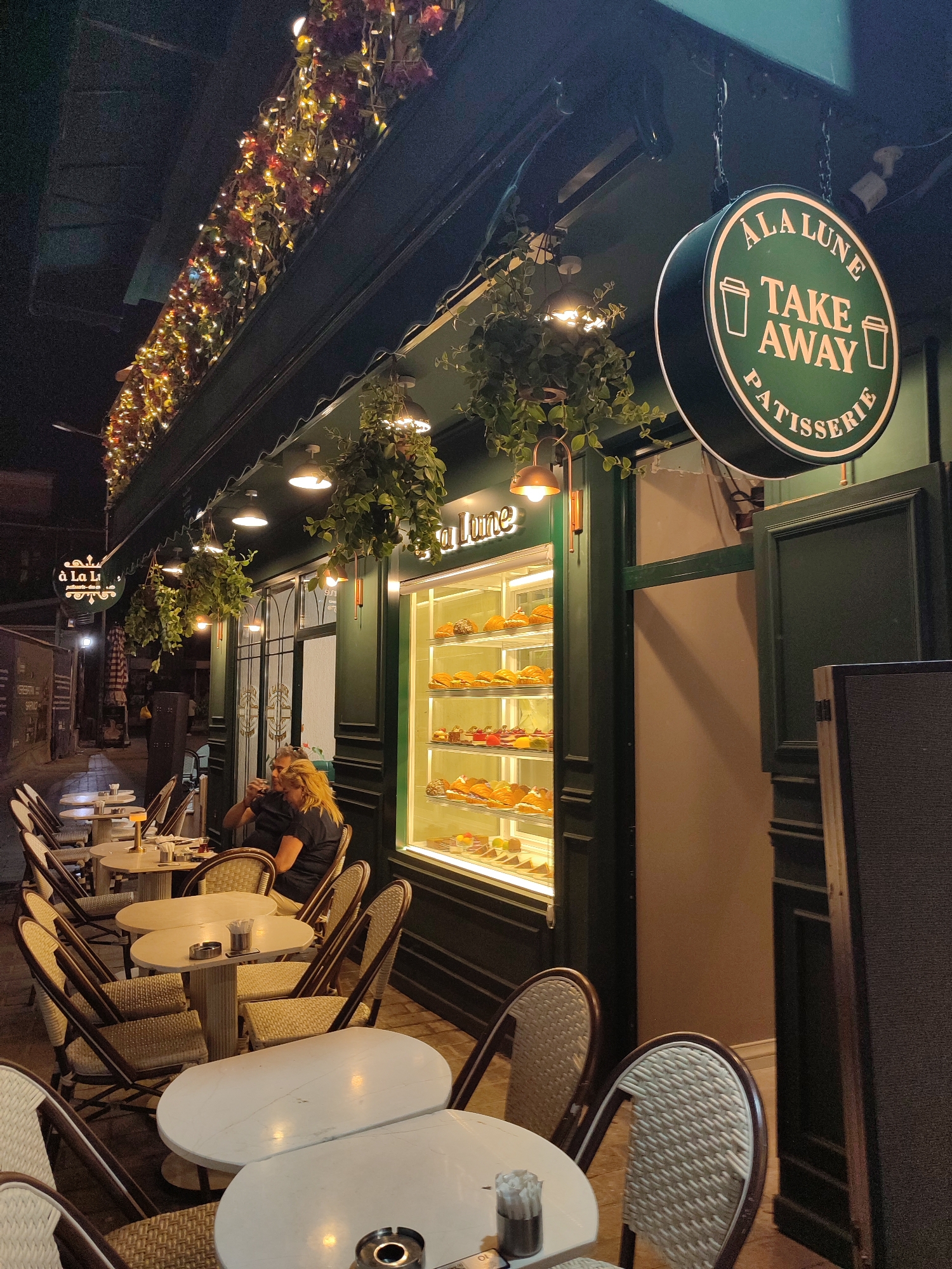 This image depicts an exterior view of a patisserie named "À la Lune" located in the Beyoğlu district of Istanbul, Turkey. The patisserie has a dark green facade with white lettering and a circular "Take Away Patisserie" sign hanging from chains. String lights and hanging plants adorn the storefront, creating a festive and inviting atmosphere. Inside, a display case showcases various pastries. Outside, several small tables with white tops and rattan chairs are set up, some occupied by patrons. One woman with blonde hair is seated at a table, holding a drink. The scene is illuminated by ambient lighting and the warm glow from the patisserie's interior, suggesting it is evening. The street outside appears cobblestoned.