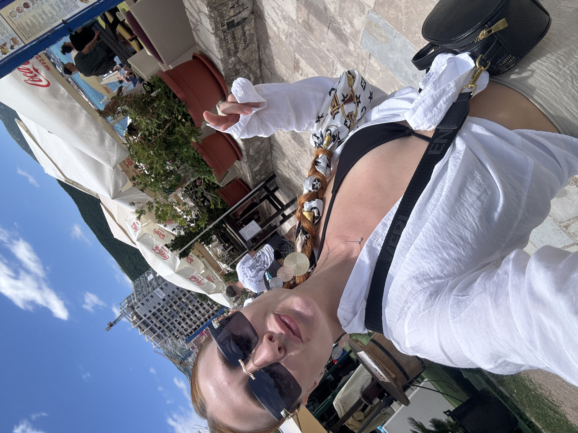 A woman with red hair braided in a pigtail is taking a selfie outdoors. She is wearing sunglasses, a white open shirt over a black bikini top, and light-colored pants. She has a black bag slung across her body. In the background, there are umbrellas, potted plants, a construction site with a crane, and a body of water. The scene appears to be a cafe or restaurant on the coast in Budva, Montenegro, with a mountain in the distance. The sky is blue with some clouds.