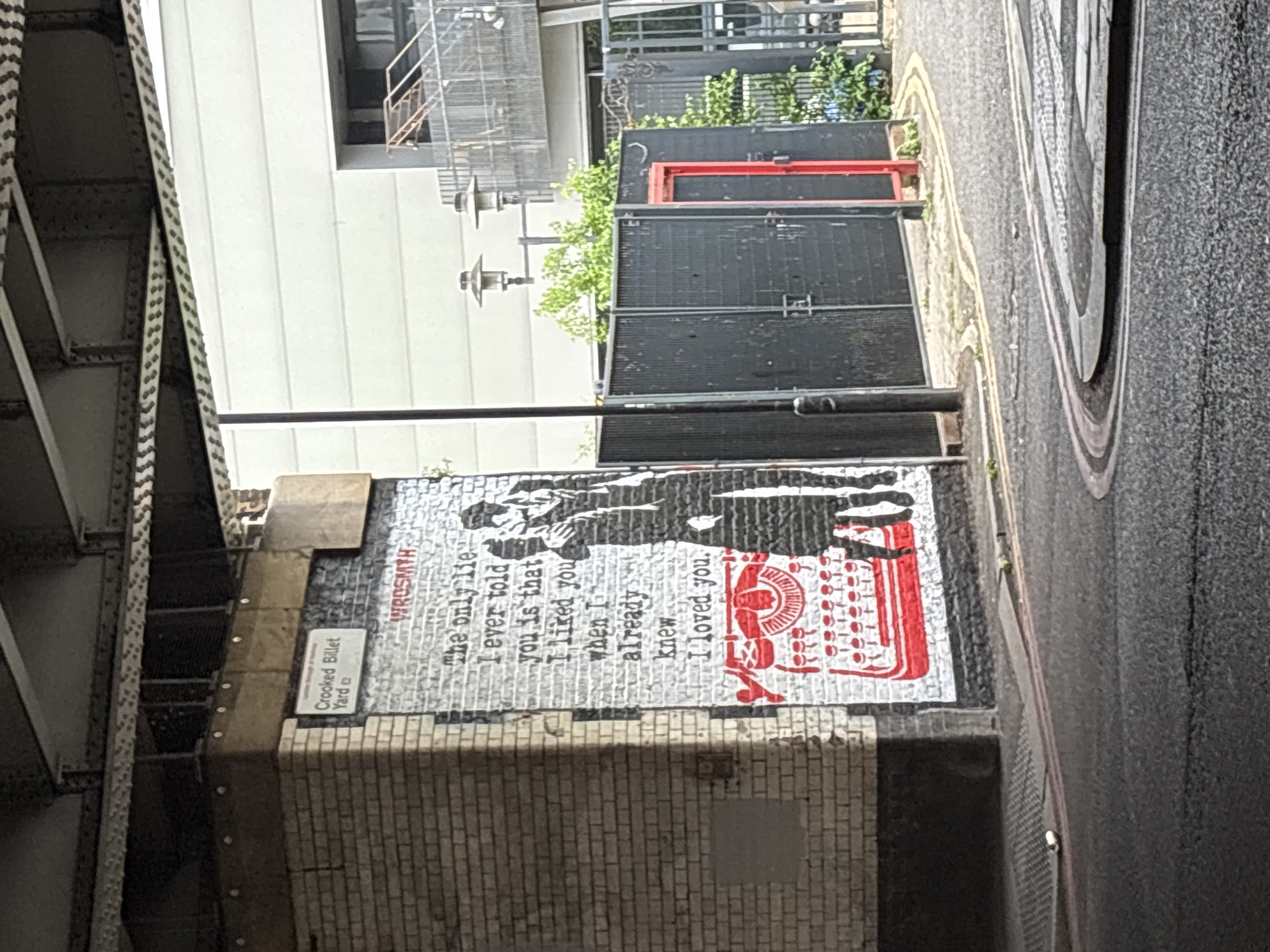 This is an outdoor, eye-level shot in an alley or narrow street in London, United Kingdom. On the right, there is a street asphalt pavement. On the left is a metal structure, possibly a bridge. Affixed to a brick wall is a sign reading "Crooked Billet Yard", under which there is a mural. The mural, attributed to WROSMTH, depicts a silhouette of a person with a typewriter and a block of text. The text of the mural reads, "The only lie I ever told you is that I liked you when I already knew I loved you.". Behind the wall with the mural is a building with an outdoor staircase.