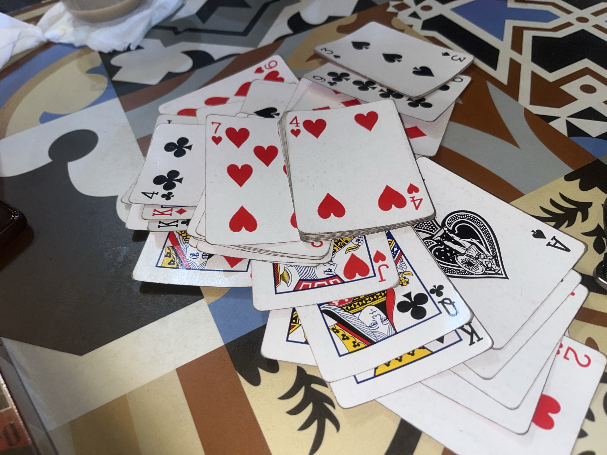 The image shows a close-up, overhead view of a pile of playing cards scattered on a patterned surface, possibly a table. The cards are a mix of suits and ranks, including Kings, Queens, Jacks, and numbered cards. Some cards are face up, revealing their designs and suits, while others are partially obscured. The surface beneath the cards features a geometric pattern with segments of brown, beige, white, black, and blue. The lighting suggests an indoor setting. The specific location in Mekar Bakti, Indonesia, is not discernible from the visual information provided in the image. There are no people or actions depicted.