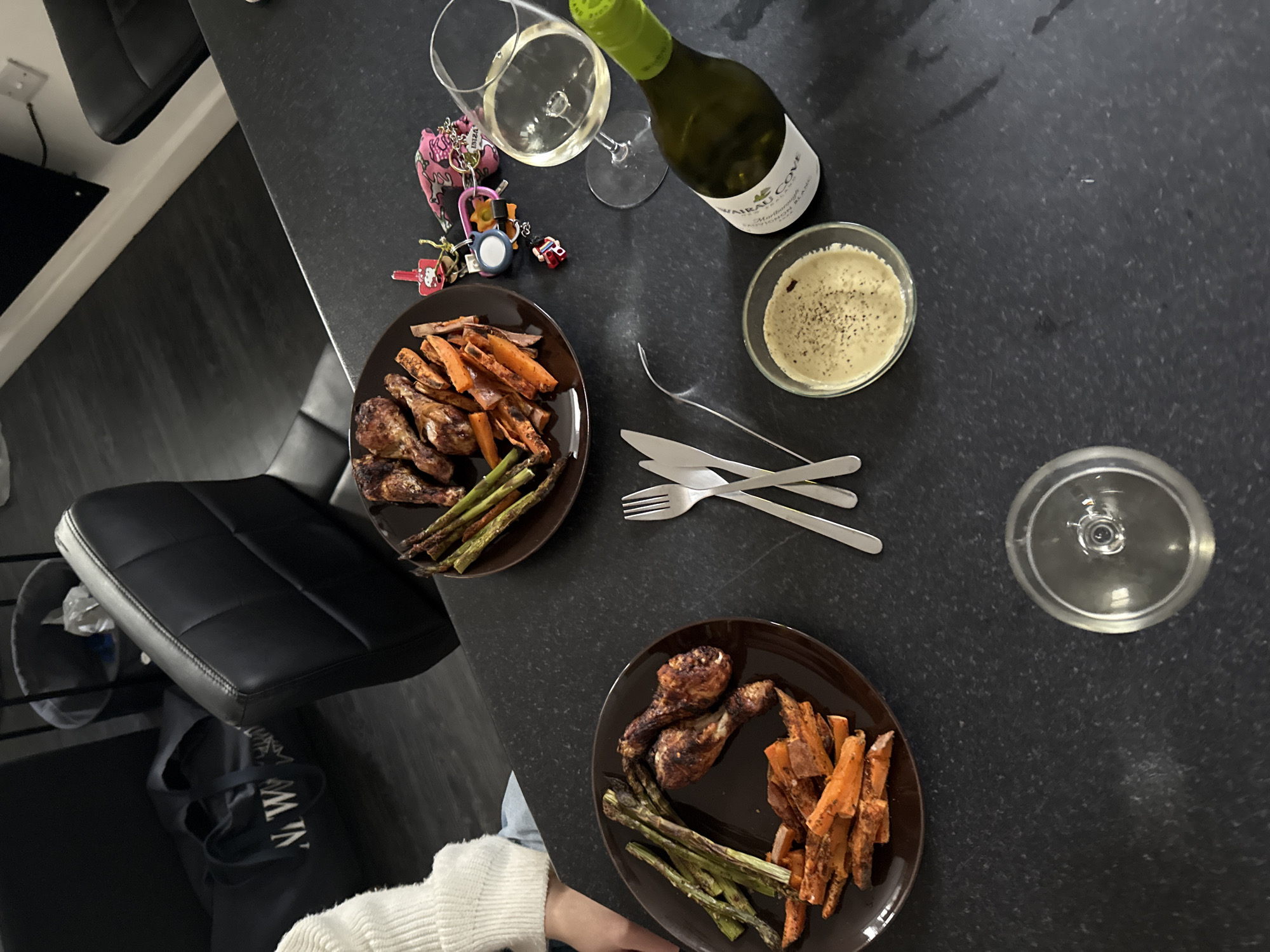This overhead shot depicts a casual dinner setting for two on a dark-speckled countertop, likely in a home in Cardiff, United Kingdom. Two dark brown plates are prominently featured, each holding a serving of roasted chicken drumsticks, roasted sweet potato fries or wedges, and grilled asparagus. Between the plates, two sets of silver forks and knives are neatly arranged.
To the right, a small clear bowl contains a light-colored creamy dip, sprinkled with black pepper. A green-labeled bottle of white wine, identifiable as "WAIRAU COVE" "MARLBOROUGH SAUVIGNON BLANC" from "NEW ZEALAND WINE," stands uncorked. One wine glass is partially filled with white wine, while a second, empty glass is positioned further to the right.
Near the wine glass, a set of keys with several keychains is visible, including a pink patterned one with the word "IBIZA" partially legible, and other small charms. In the lower left foreground, a person's hand and arm, wearing a white knitted long-sleeved top, are visible, suggesting they are seated at the counter. The background shows parts of dark chairs or bar stools and a dark floor, indicating an indoor dining area. The overall scene suggests a relaxed and intimate evening meal.