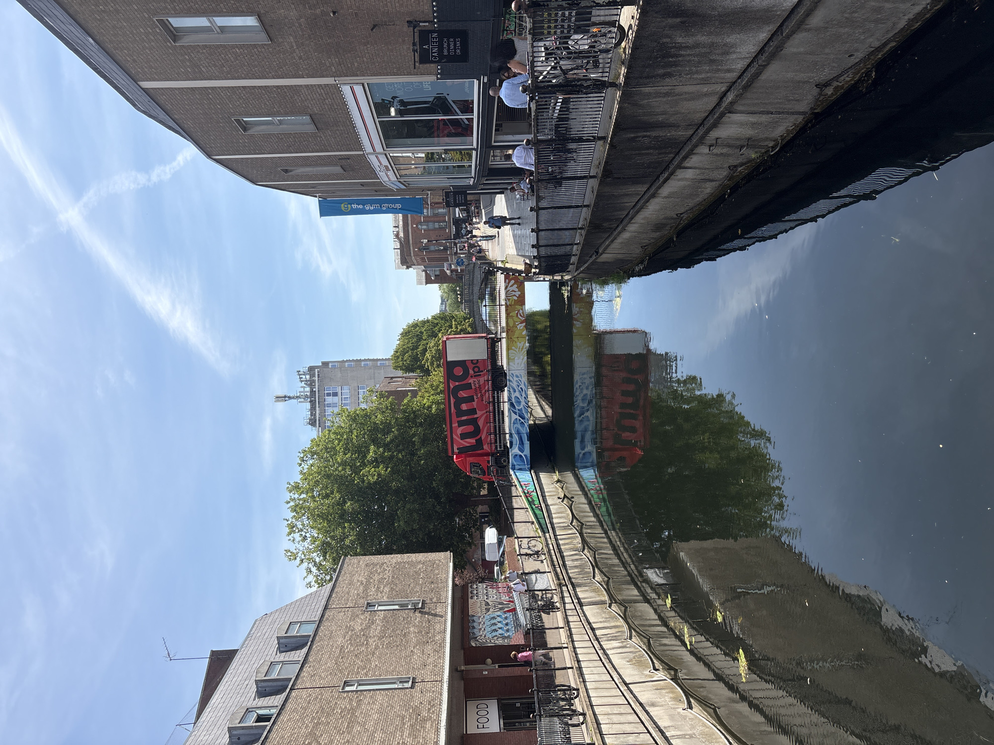 The image captures a canal in an urban environment. On the left bank of the canal, a brick building with a sloped roof and dormer windows is visible, featuring a sign that reads "FOOD". Further along the left bank, a colorful mural adorns the wall. People are seen walking and cycling along the canal path.
A bright red truck with "Luma" written on its side is positioned on a bridge crossing the canal. The bridge itself is decorated with a colorful mural of flowers. Across the canal, on the right bank, a brick building with large windows houses a gym, indicated by a blue banner that reads "the gym group". Another establishment, "A CANTEEN", is also present, with signage for "BRUNCH", "DINNER", and "DRINKS". More people are seated at outdoor tables on the right bank.
The canal water reflects the surrounding buildings, trees, and the sky, which is blue with wispy clouds and contrails from an aircraft forming an 'X' shape. The scene suggests a bustling city scene on a sunny day. The canal is lined with concrete walls and metal railings.