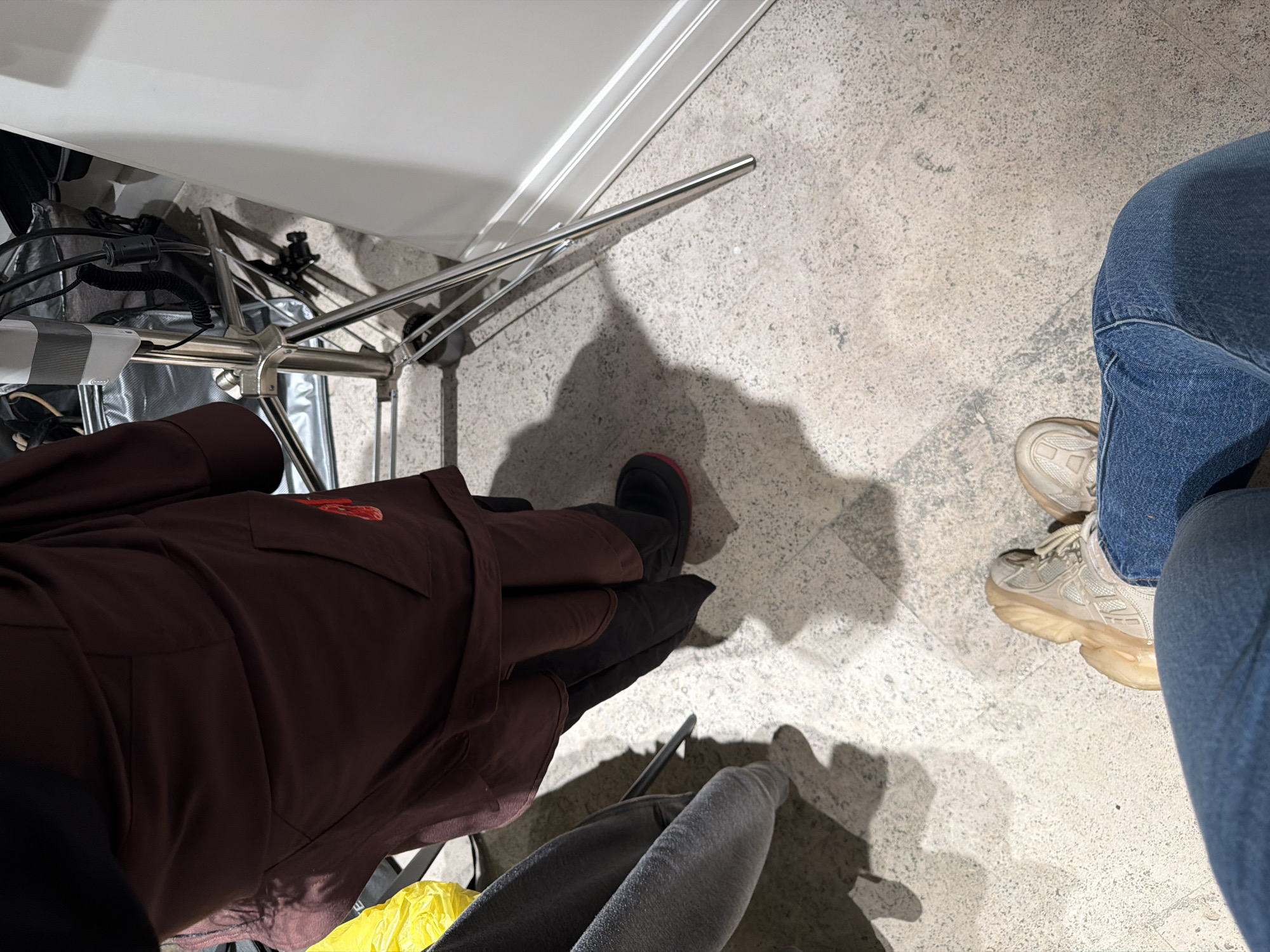 The image shows a view from above, seemingly from a seated position. To the left, a person is wearing a dark brown, long-sleeved garment and black shoes with a red sole visible. Part of the chair is visible with silver metal legs. Another person, wearing blue jeans and light-colored sneakers, is positioned to the right. The scene takes place on a grey tiled floor, and part of a yellow plastic bag and a grey sweater is visible. Without further context, it is not possible to determine if the image was taken in Yerevan, Armenia.