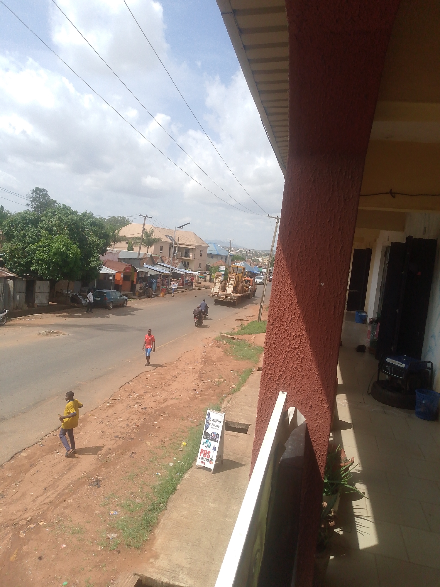 The image shows a street scene in an urban area. There is a paved road with a person walking on the shoulder. Buildings are visible on both sides of the street. A man walks in the street, wearing a yellow shirt and dark pants. Construction equipment can be seen in the distance on the road. A portable sign is near the sidewalk, advertising a "POS Available Here". Power lines stretch across the sky. The photo is taken from an elevated position, possibly a balcony or upper floor of a building. A portion of the building's architecture is visible in the foreground.