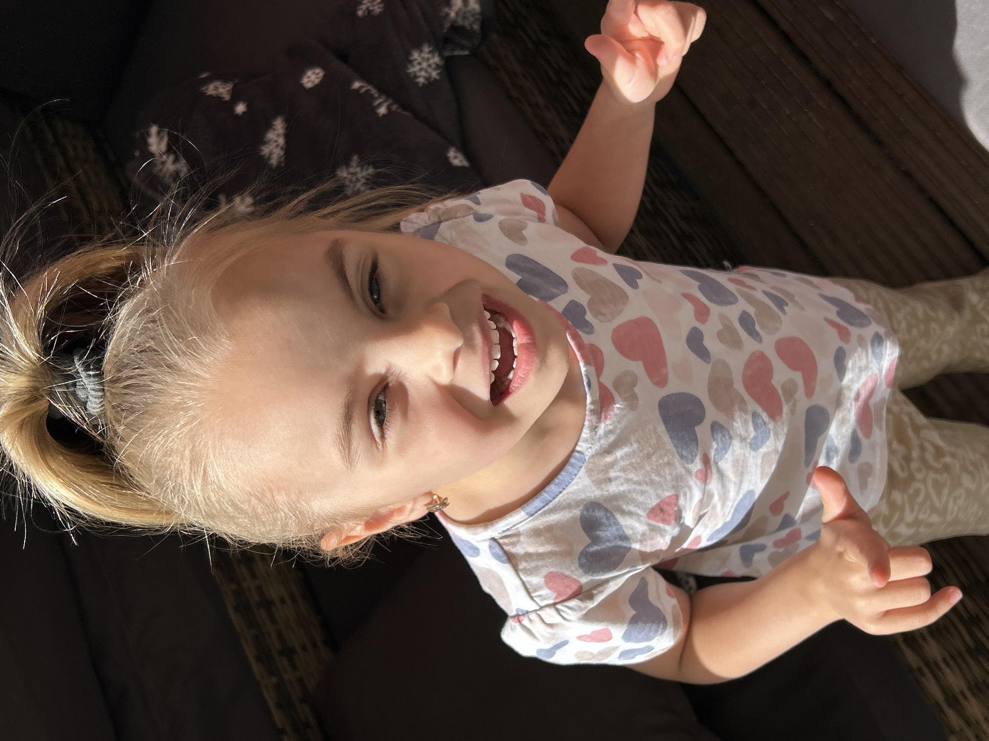 A young girl with blonde hair tied in a ponytail is looking up at the camera. She is wearing a t-shirt with a heart pattern and light-colored pants. She has a big smile on her face, showing her teeth. The image appears to be taken outdoors, with natural light casting shadows. The background consists of dark furniture and wooden planks, suggesting an outdoor seating area or deck. The specific location in Waldbröl, Germany is not discernible from the visual information provided.
