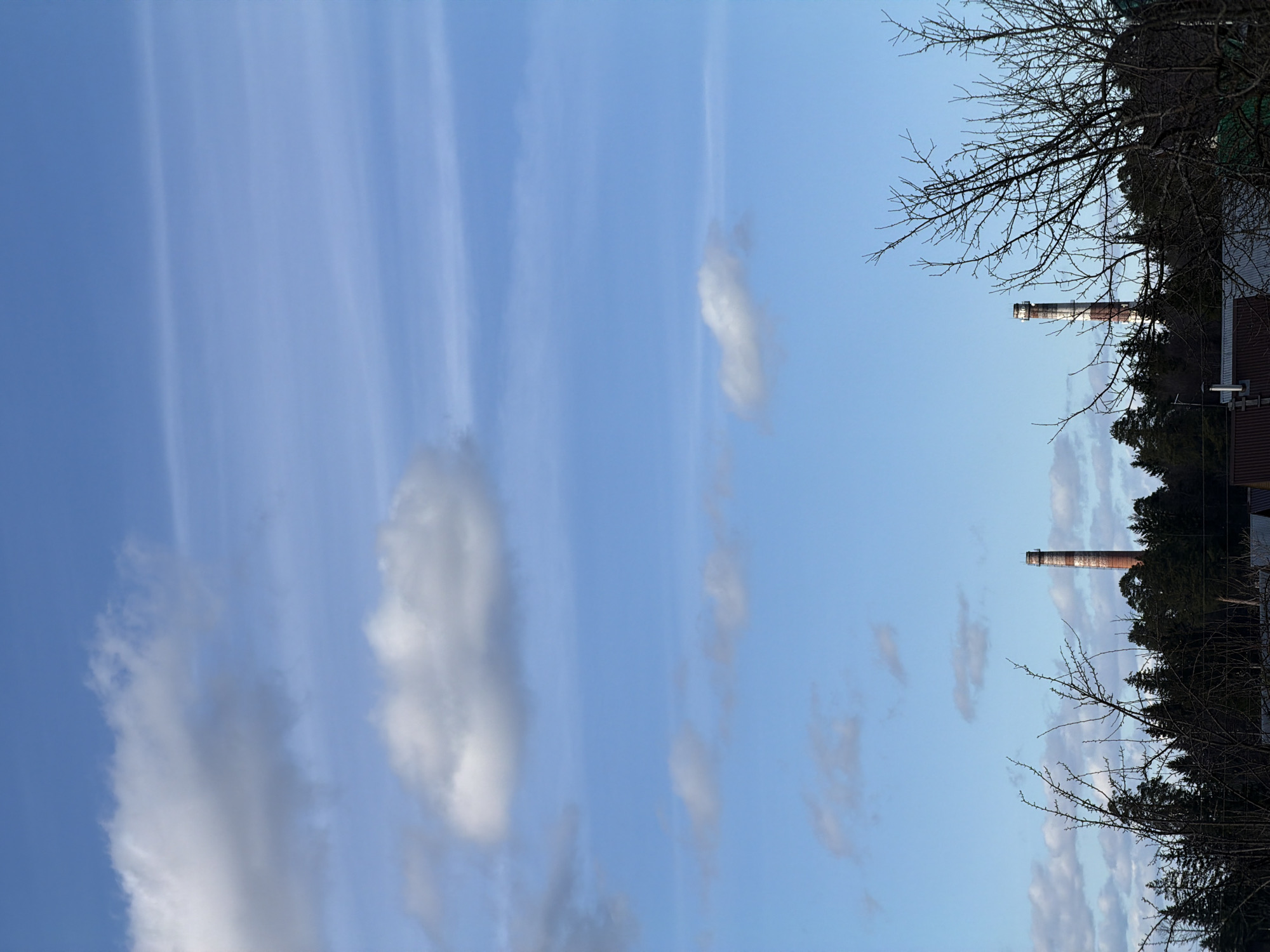 The image shows a view of the sky with scattered clouds and two industrial chimneys in Chaikovsky, Russia. The chimneys are made of brick and feature alternating red and white bands. They are positioned behind a line of trees and vegetation, with bare tree branches visible in the foreground. A portion of a building's roof, possibly a house or industrial structure, can be seen near the lower chimney. The sky is predominantly blue with wispy clouds, suggesting fair weather.