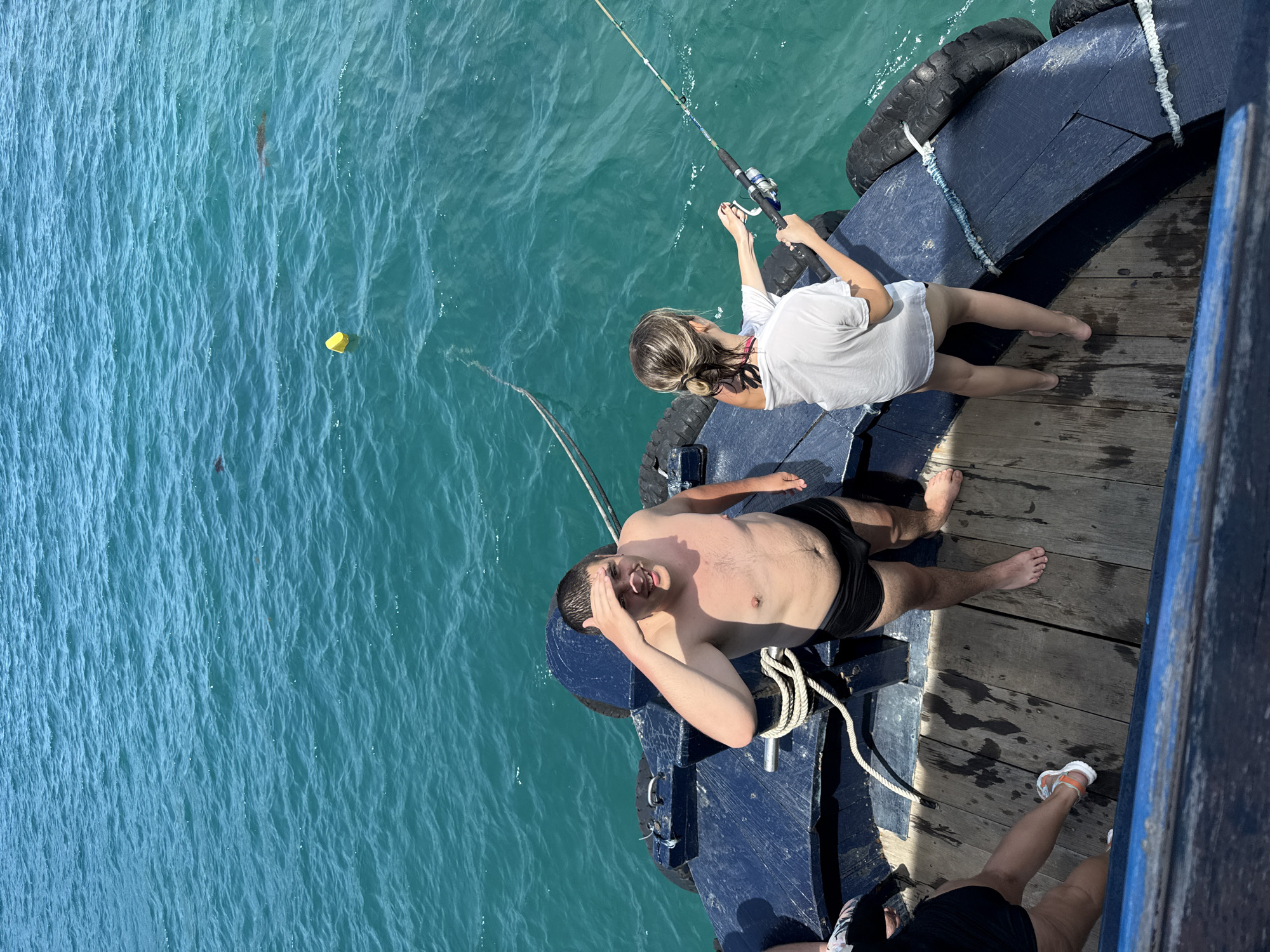Captured from an elevated angle, the scene unfolds on a pier or boat deck in what appears to be a coastal environment. The turquoise-hued water stretches out beside the structure. A fair-skinned woman in a white top and shorts is engaged in fishing, holding a rod with both hands. Adjacent to her, a man, shirtless and wearing black shorts, is lying down, his hand shielding his eyes from the sun. Another individual's legs are partially visible in the lower right corner, clad in black shorts and white sandals. A yellow object floats in the water. The scene is situated in Nha Trang Ward, Vietnam.