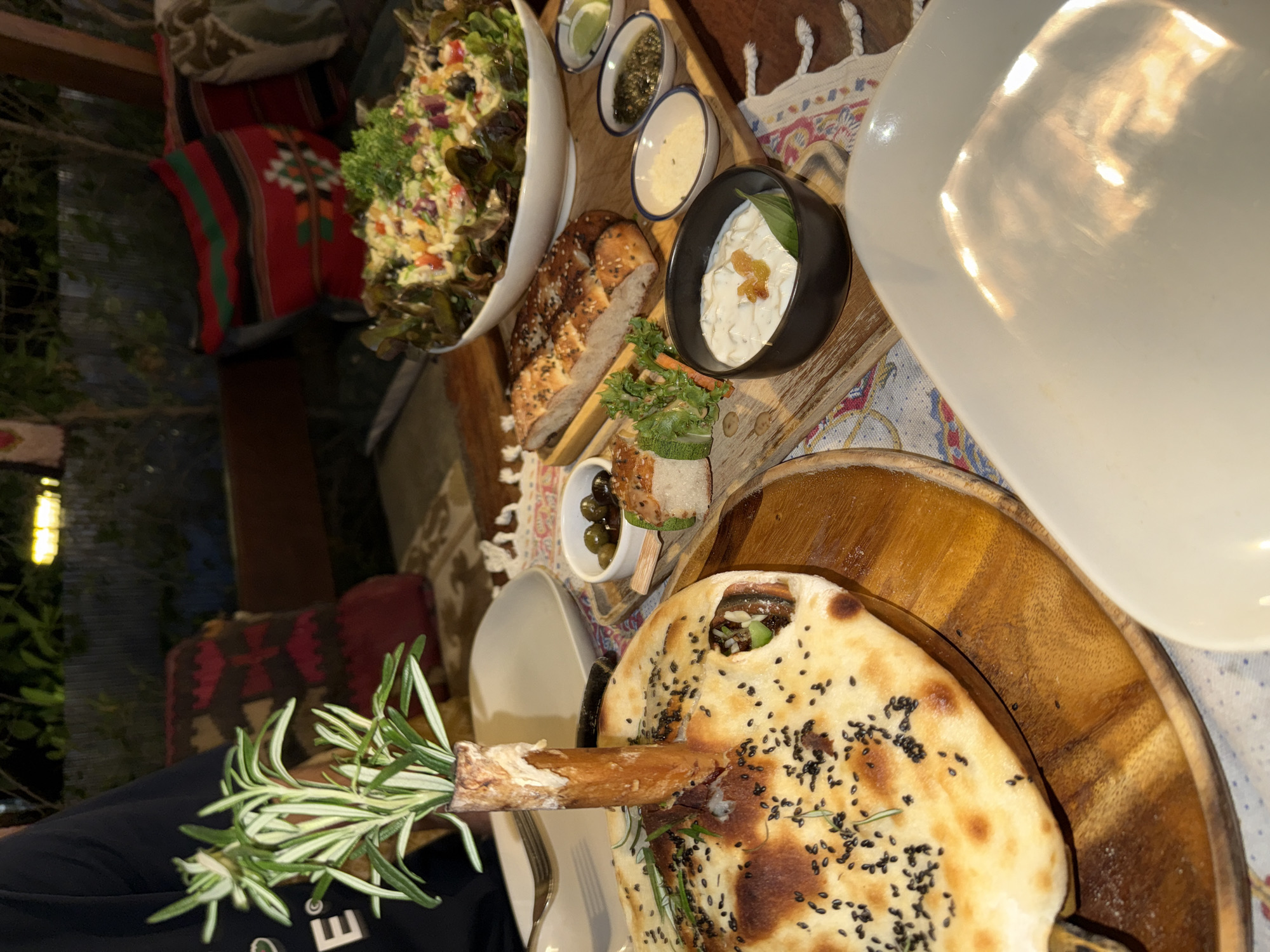 The image depicts an outdoor dining scene in what appears to be Baan Hin Kong, Thailand. A wooden table holds various dishes arranged for a meal. A large salad in a white bowl and several smaller bowls containing condiments or dips are visible. Bread, including naan-like flatbread sprinkled with black seeds, is also presented. One piece of bread has a sprig of rosemary protruding from it. A large white plate occupies the upper right of the frame, obscuring part of the scene. Pillows with Thai patterns sit on seats behind the table. The lighting suggests it's either late afternoon or evening. A person wearing a black shirt with white lettering is partially visible in the lower left corner.
