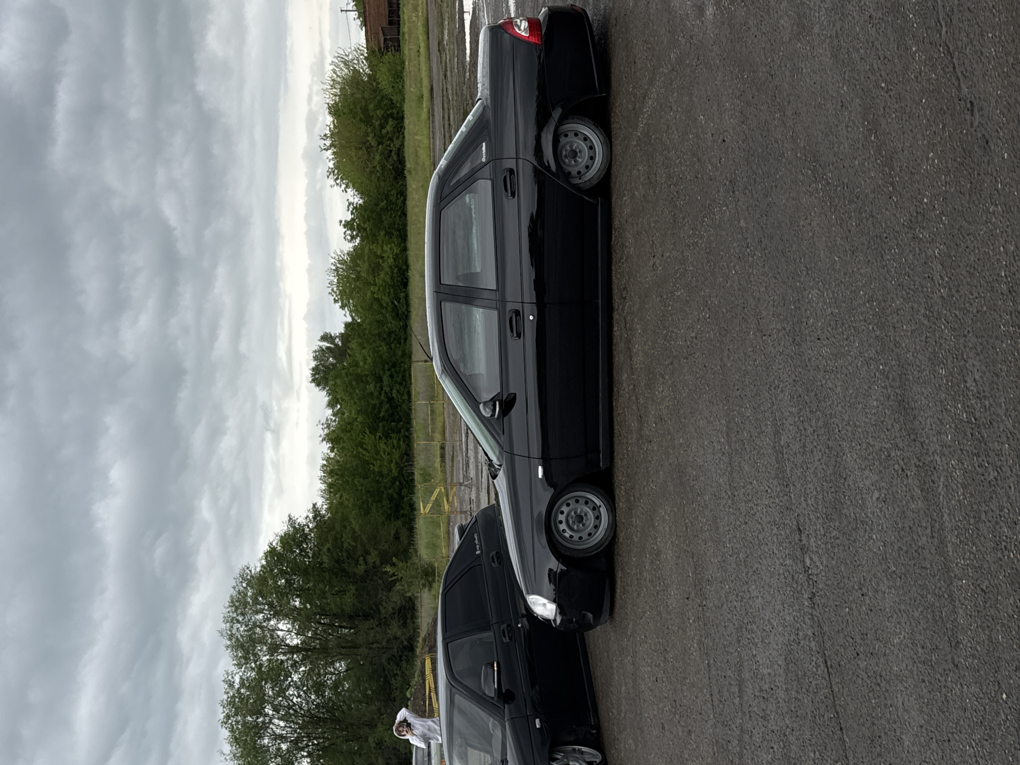 The image shows two black Lada Priora sedans parked on an asphalt surface. Trees line the background, with an industrial structure faintly visible behind them. The sky is overcast. A person in a white hooded garment stands near the first Lada. The location is potentially Pavlovskaya, Russia.