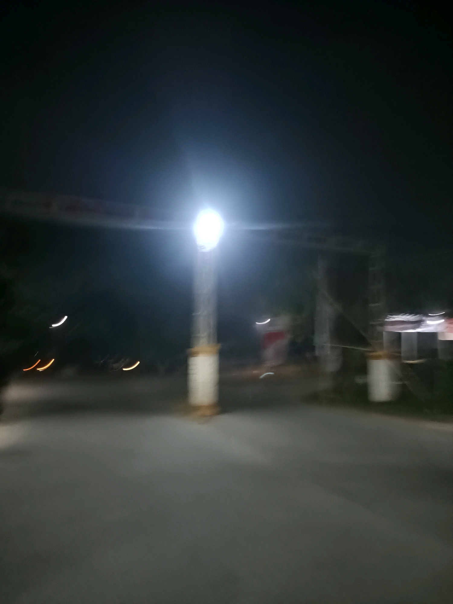 The image depicts a blurred nighttime scene on a paved road, likely an entryway or a toll booth area in Telagaasih, Indonesia. A prominent streetlamp illuminates the scene with a bright, diffused light. In the background, there appears to be some sort of structure or building, also partially lit and blurred. Several curved light streaks, possibly from vehicle headlights or taillights, are visible, indicating movement. The overall impression is one of motion and low light conditions, common in urban or semi-urban settings at night.