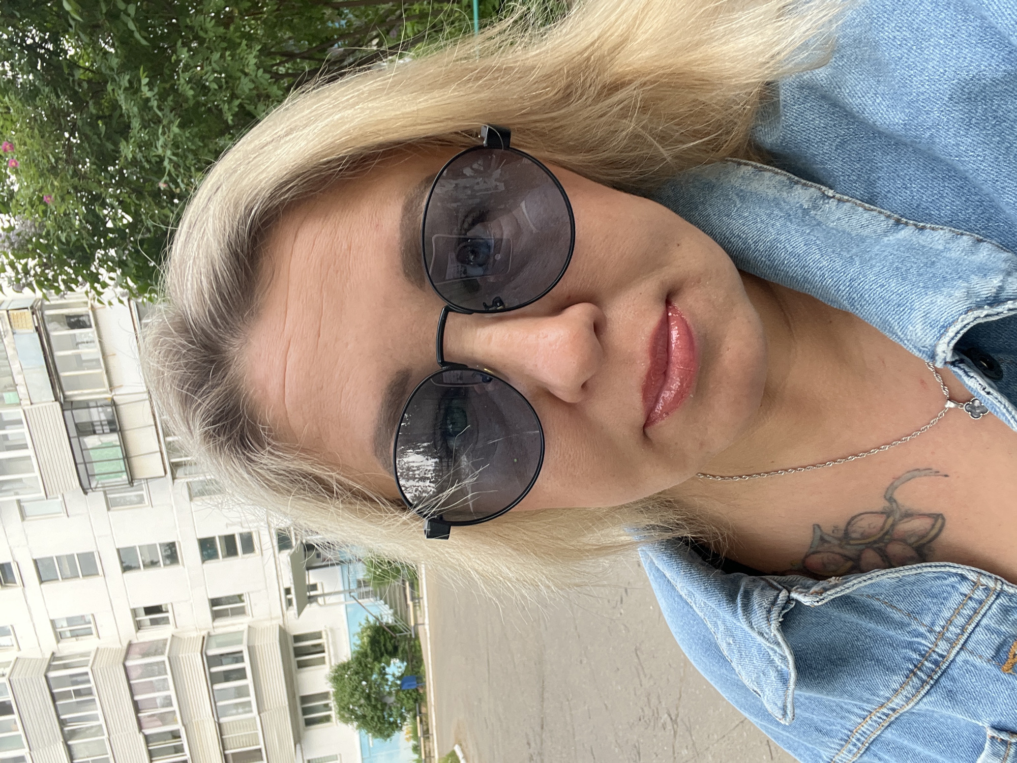 A selfie of a fair-skinned woman with blonde hair wearing round, black sunglasses and a light blue denim jacket. She also has a silver necklace and a tattoo on her upper chest. The background reveals an urban environment with a white apartment building and green trees, possibly in Chaikovsky, Russia. The reflection in her sunglasses shows additional details of the surroundings, but specifics are unclear.