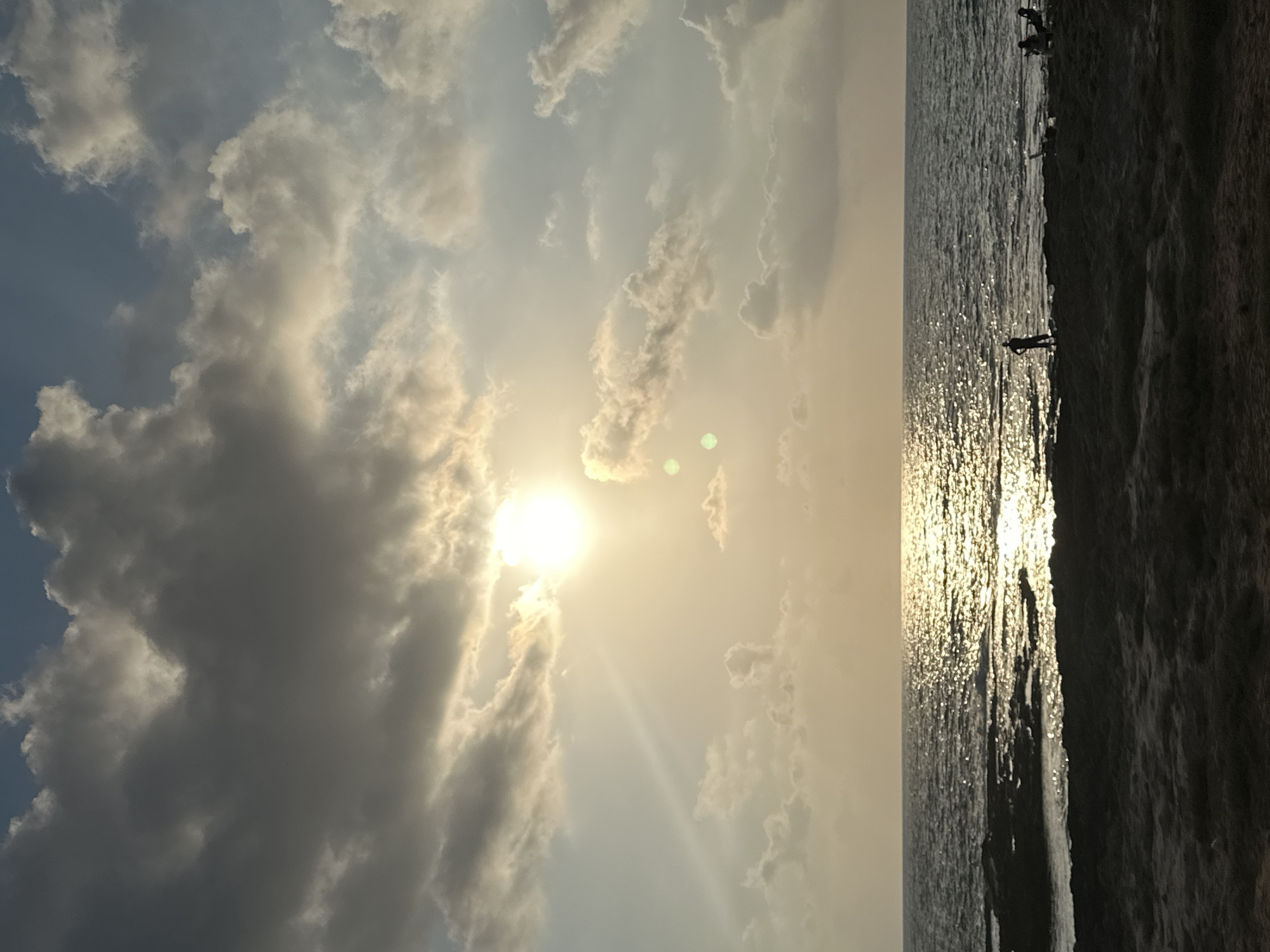 A sunset over the Mediterranean Sea casts a golden glow on the water and the sky. Dark clouds are scattered across the sky, with the sun peeking through them. The waves are gently lapping at the shore, where a few people are visible. The scene is captured from a beach, with sand in the foreground. The location is likely the promenade in Nahariyya, Israel, given the commonality of such sunset views along the coastline there.
