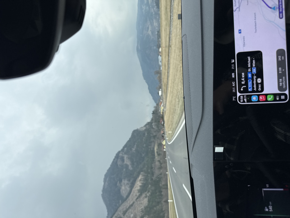 The image provides a forward-facing view from inside a vehicle, depicting a rural road scene in Teufenbach-Katsch, Austria.
**Vehicle Interior:** The lower portion of the frame shows a dark-colored vehicle dashboard, featuring a large, integrated digital display. This screen displays a navigation interface showing a route with "8,4 KM" to the next turn, listing destinations like St. Michael, Judenburg, Graz, and Wien. The map area clearly identifies "Teufenbach-Katsch" as a nearby geographical feature. Additional digital readouts include the time "15:58", external temperature "7°C", cabin A/C setting "20.5 A/C" with an auto mode, target cabin temperature "21.5°", full LTE signal, estimated arrival time "18:18", travel duration "2:20", and remaining distance "242 KM". On the left side of the display, a gauge indicates a "540 KM" range and "1/2" fuel tank level. A portion of the vehicle's rearview mirror housing is visible at the top center of the image. The windshield shows minor dust or streaks.
**Exterior Environment:** A two-lane asphalt road, marked with white dashed center lines and solid white shoulder lines, extends into the distance, curving gently to the right. White roadside markers are visible along the right shoulder. To the left of the road, a large, steep mountain rises, characterized by exposed rock faces and areas of sparse, dark-colored vegetation. To the right of the road, a wide, flat expanse of dry, yellowish-brown grass is visible, across which a set of railway tracks runs parallel to the road. Further right, a cluster of houses with reddish-orange roofs is situated along the base of distant hills. The sky is uniformly overcast and grey, suggesting cloudy weather. No individuals are visible in the scene.