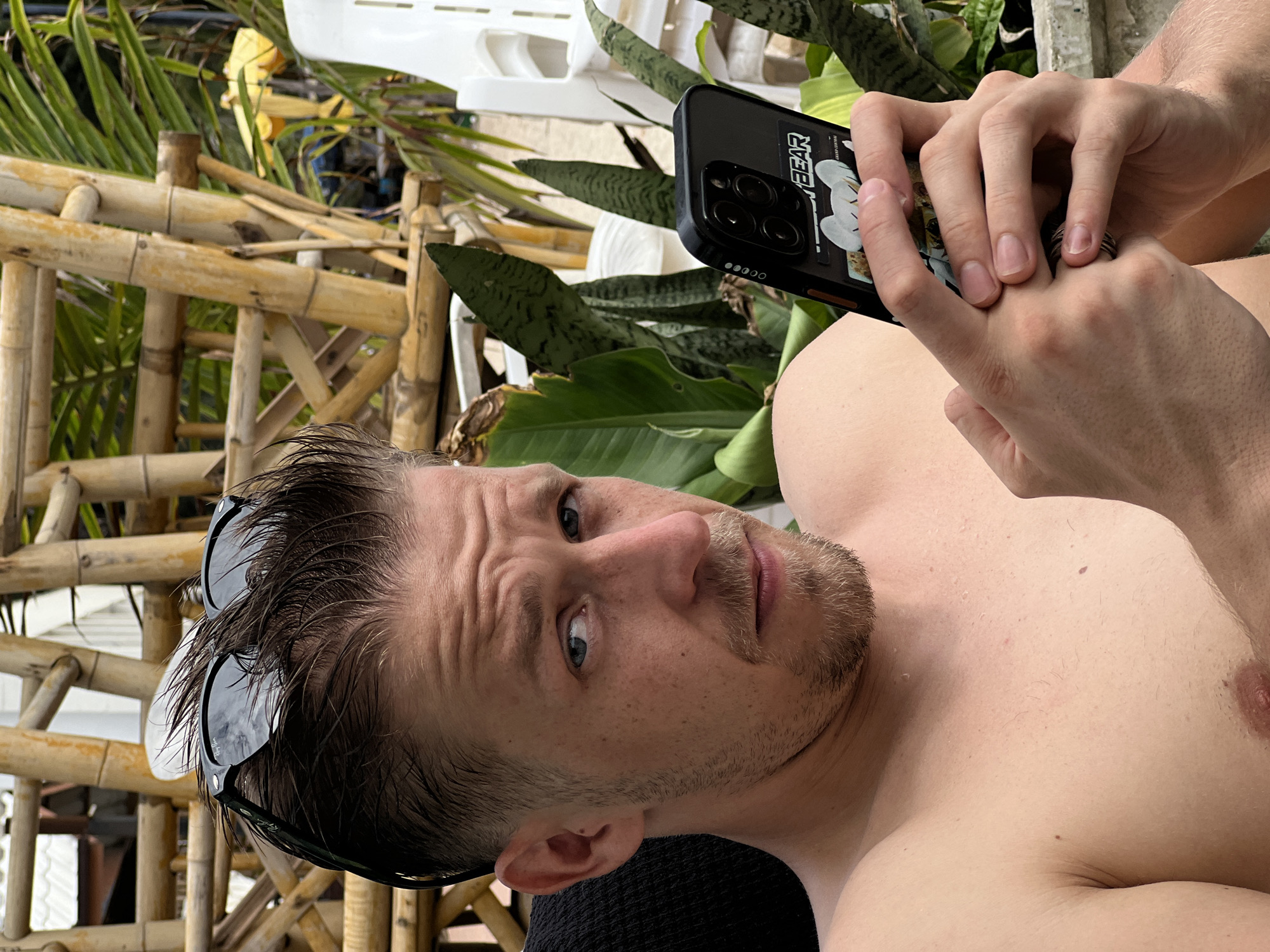 A shirtless man with fair skin and light brown hair is holding a smartphone in his right hand and looking at the camera. He is wearing sunglasses on top of his head. The man has a light beard and appears to be outdoors, with a background of bamboo structures and green foliage. The setting suggests a tropical or resort-like environment.