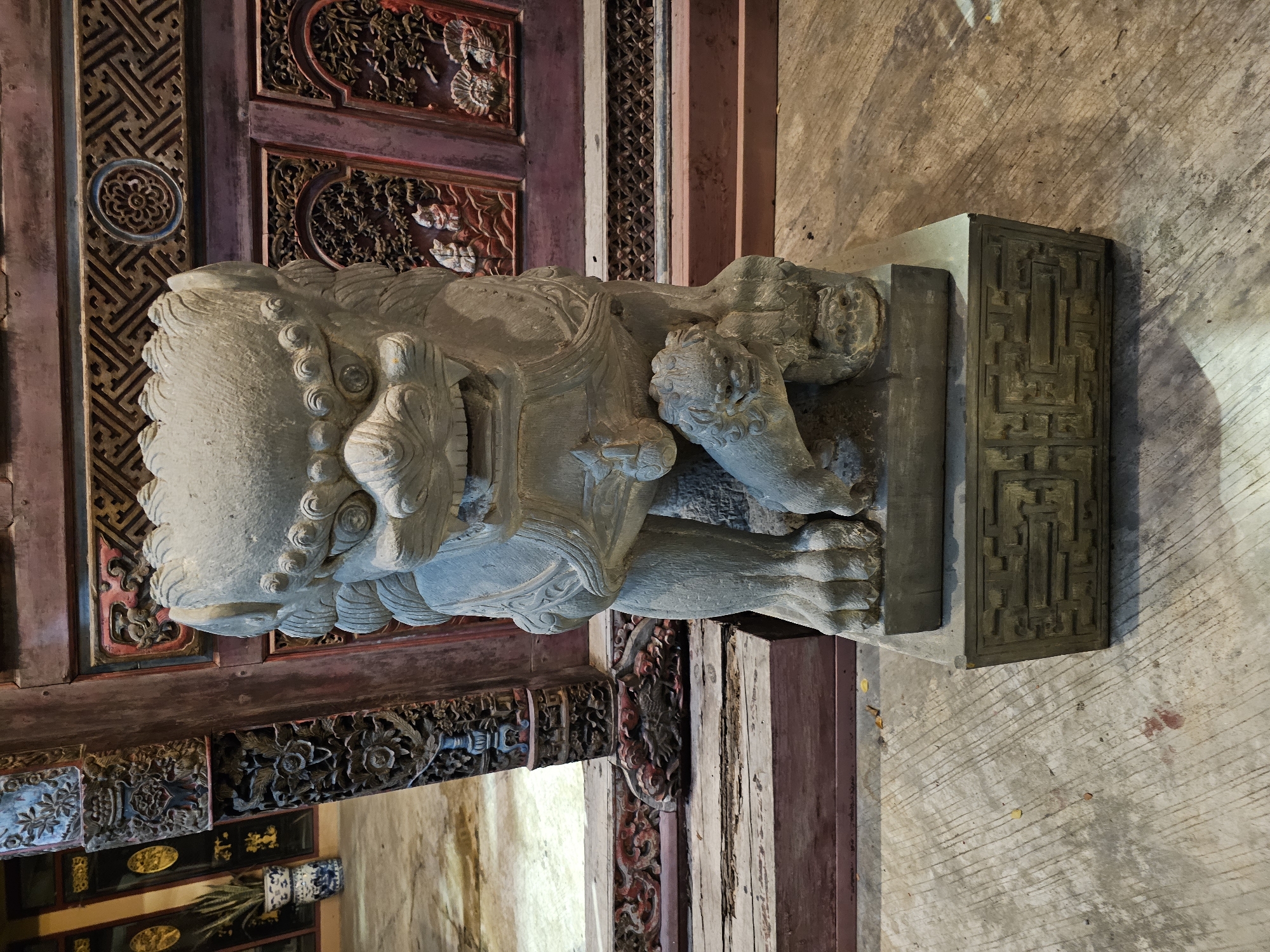 A weathered stone sculpture of a lion and a cub stands on a decorative stone base in front of an ornate wooden structure. The lion sculpture is positioned centrally, with its mouth open in a fierce expression and its body leaning forward. A smaller lion cub is positioned beneath the larger lion's front paw. The wooden structure behind is intricately carved with floral and geometric patterns and painted in dark red and gold accents. The ground is a textured concrete surface. The scene appears to be indoors or in a covered area, possibly a temple or traditional building, with soft lighting. The location is identified as Bangkok, Thailand.