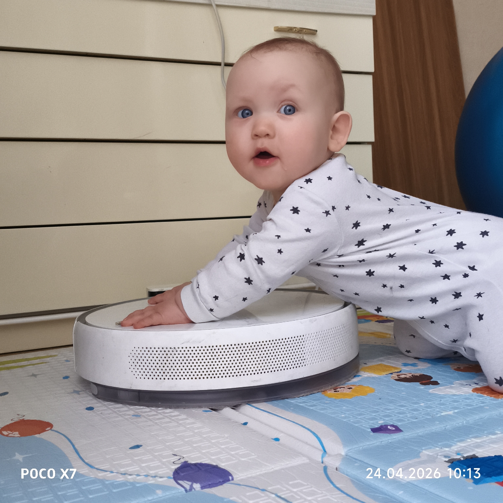 A baby wearing a white onesie with black stars is crawling on a blue and white patterned play mat. The baby has blue eyes and is looking at the camera with an open mouth. The baby's hand is resting on a white robot vacuum. The background includes a white dresser with drawers and a blue exercise ball. The date "24.04.2026 10:13" and the text "POCO X7" are visible in the corners of the image. There is no information to indicate this photo was taken in the city of Chaikovsky, Russia.
