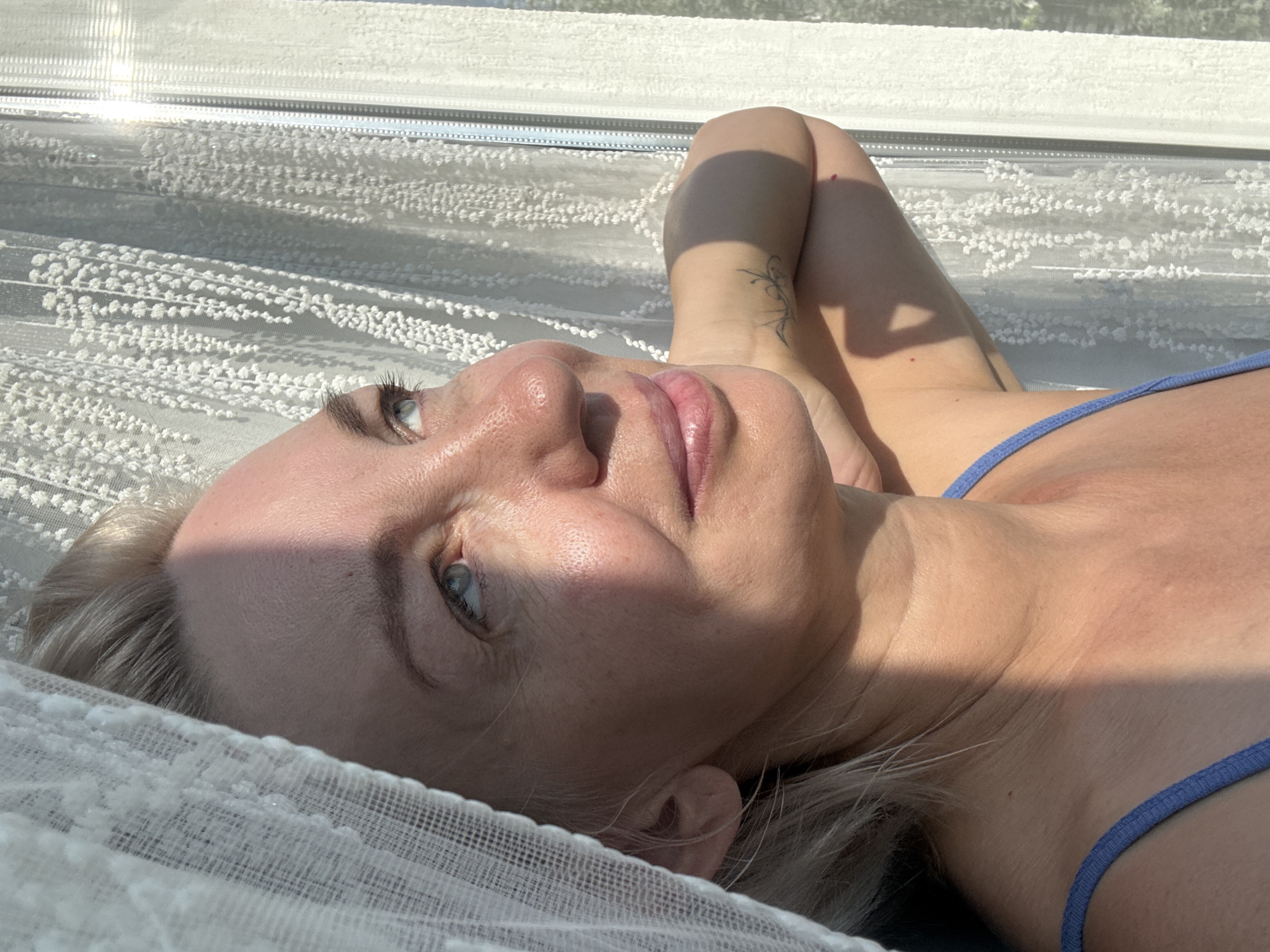 A blonde woman with light blue eyes and fair skin, in her 40s or 50s, is positioned in the foreground of the image. She is looking upwards and to the right, with her head tilted back and her arms behind her head, partially obscured by a white sheer curtain on the left. Sunlight casts a strong shadow across the left side of her face, highlighting freckles and fine lines. She is wearing a blue tank top. The background features a white lace curtain hanging in front of a window, with sunlight streaming through it, creating a bright, airy ambiance. The scene appears to be indoors, near a window, possibly in a residential setting. There is no specific information or visual cues in the image that suggest the location is the city of Sochi, Russia.