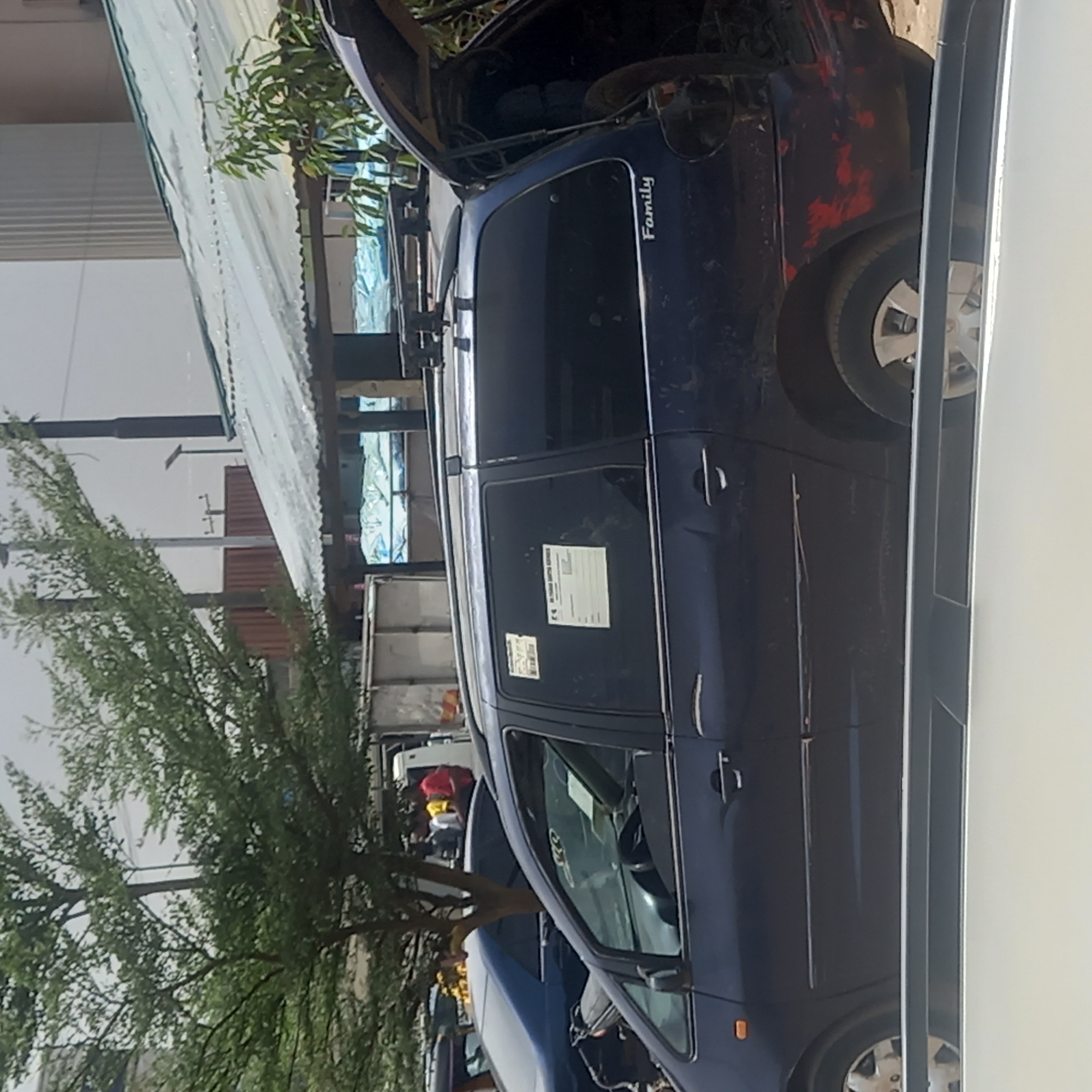 The image captures a dark blue minivan or MPV parked outdoors in Dadi Riba, Nigeria, under bright daylight. The vehicle, positioned prominently in the frame, appears to have its hood or rear boot open, possibly indicating maintenance or loading activity. A visible roof rack runs along the top of the car.
Distinctive features on the car include a white sticker with the word "Family" printed vertically on the rear passenger-side window. Another rectangular paper document, likely a permit or information sticker, is affixed to the front passenger-side window, though its text is illegible. The car's body shows some wear, with patches of red visible on the rear fender and door, possibly from damage or an underlying paint layer. The rear wheel on the visible side is missing its hubcap.
The background features several elements typical of a semi-urban or commercial area. Lush green trees are visible to the left, and various buildings, including one with a corrugated metal roof, stretch across the scene. Utility poles and power lines are also present against the clear sky. Further back, other vehicles, including what appears to be a truck, and indistinct figures can be seen, suggesting a bustling environment. The strong sunlight indicates it is daytime with clear weather.