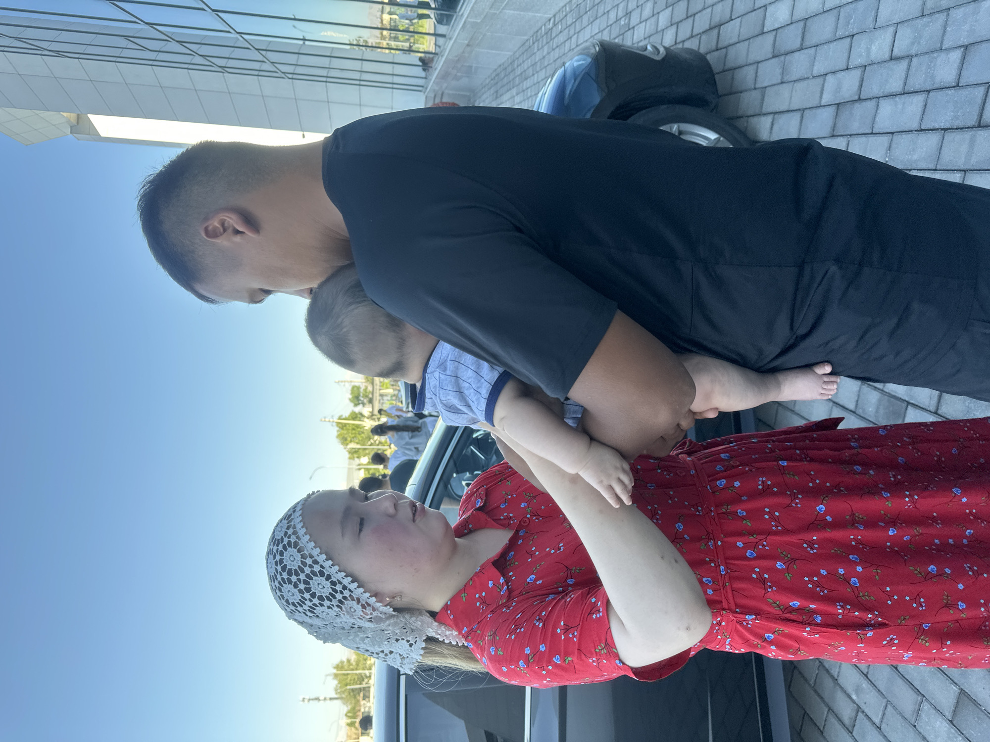 The image shows a family in what appears to be Manas, Kyrgyzstan. A man in a black t-shirt is holding a baby. A woman in a red floral dress and a white lace headscarf is looking up at the man and baby. The scene is outdoors, with a clear blue sky and some trees in the background. The ground is paved with grey bricks. A black car is partially visible. The overall impression is a warm and sunny day, and the subjects appear to be happy and connected. The time of day seems to be daytime, likely afternoon, given the brightness of the sky and the shadows.