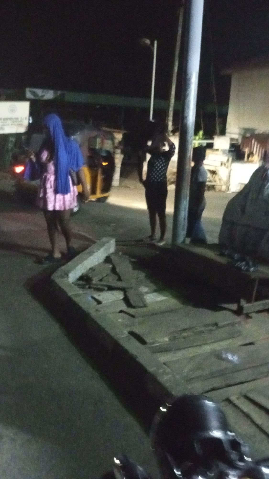This nighttime image captures a street scene in Lokoja, Nigeria, under dim artificial lighting. The photograph appears to be taken from a low angle, possibly from a motorcycle, with a dark, metallic object resembling a handlebar visible in the immediate foreground.
Several individuals are present in the frame. On the left, a person wearing a pink dress and a blue head covering stands, partially obscuring a yellow and black tricycle (Keke Napep) whose red taillight is visible. In the mid-ground, a tall, light-colored pole stands prominently, partially obscuring another person dressed in a dark, patterned top and dark shorts or a skirt, with their hands raised towards their head. Further to the right, a third person is partially visible, wearing a dark top and lighter trousers, standing beside what appears to be stacked items or bags.
In the lower mid-ground, a low concrete structure is visible, covered with several wooden planks. A blurry white sign with indistinct dark text can be seen on the far left. The background shows faint lights and the vague outlines of buildings, suggesting an urban or semi-urban setting. The scene conveys a sense of a typical evening or late-night street environment, with people gathered in a public space, though no specific activity is clearly discernible.