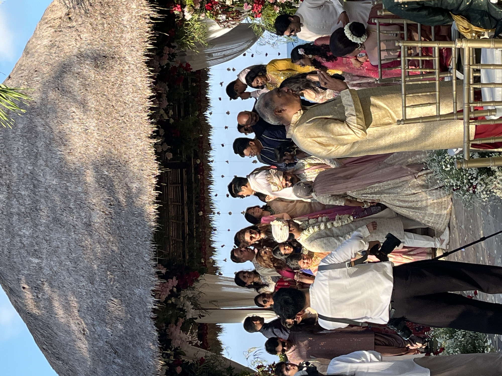 In Kuta Selatan, Indonesia, under a thatched-roof pavilion adorned with floral arrangements, a wedding ceremony is taking place. A group of people, dressed in traditional Indian attire, are gathered. The bride and groom are visible, with the groom wearing a cream-colored sherwani and a turban, and the bride in a light pink lehenga. Several guests are observing the ceremony, with some dressed in vibrant colors. A photographer is positioned in the foreground, equipped with a professional camera and tripod, documenting the event. The background reveals glimpses of the ocean, suggesting a coastal setting.