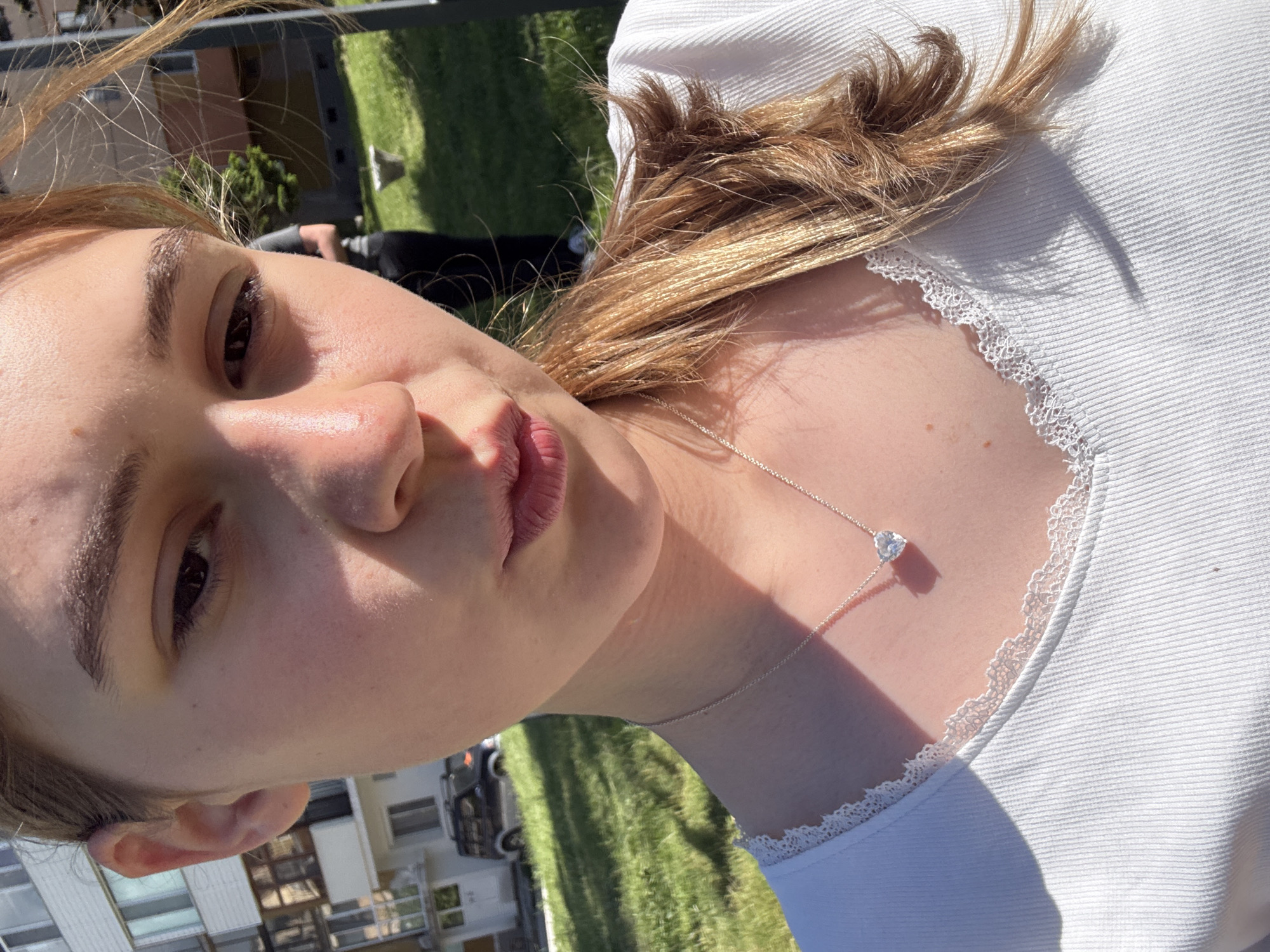The image is a selfie taken by a fair-skinned individual with brown hair and eyes. They are wearing a white top with lace trim and a silver necklace with a heart-shaped pendant. The background shows a grassy area and a building. The building appears to be residential. Based on the background details, it is possibly taken in the city of Panagyurski kolonii, Bulgaria.