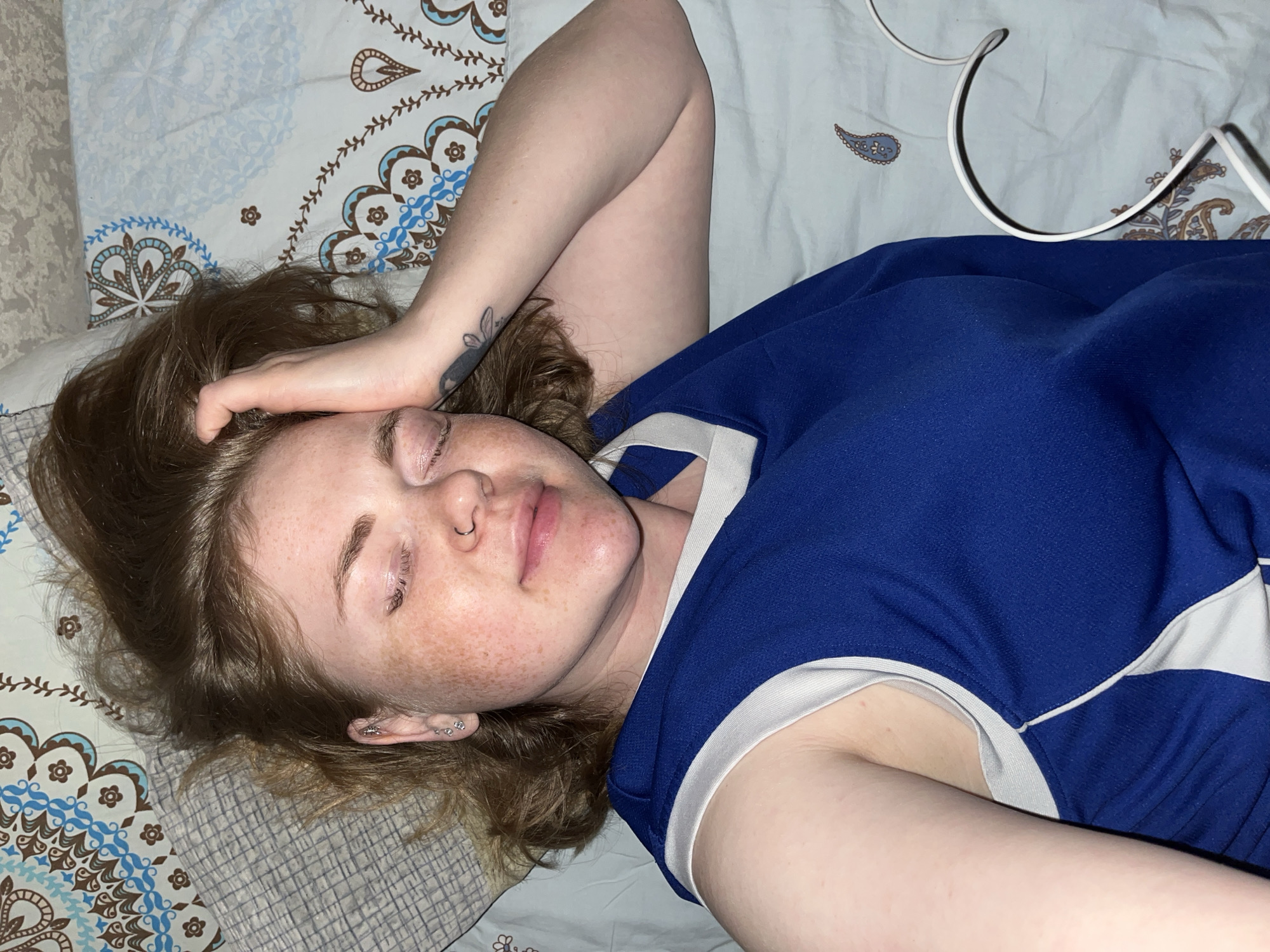 A fair-skinned person with auburn hair is lying on a bed. They are wearing a blue and white top. The person's eyes are closed, and they have freckles on their face. One arm is bent and raised, with the hand resting on the person's head. The person has a nose piercing and multiple earrings. There is a tattoo on the inner forearm of the raised arm. The bed is covered with a patterned sheet and pillow. A white cable is partially visible in the upper right corner. There is no indication of this image being from Chaikovsky, Russia.