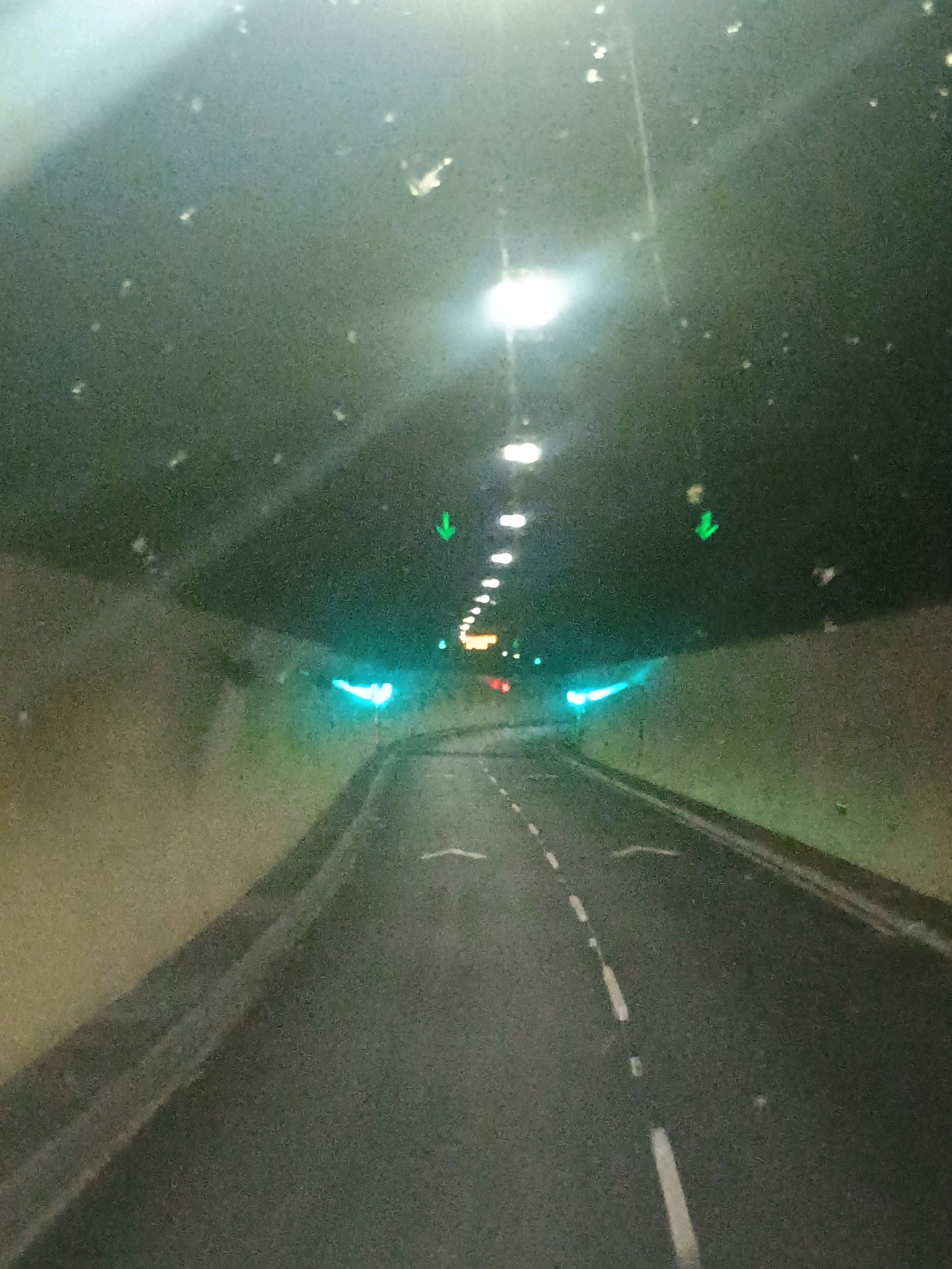 The image shows a view from a vehicle traveling through a tunnel at night. The tunnel has a dark, curved ceiling with rows of bright lights. The walls of the tunnel are a light brown color. The road ahead is asphalt with white lane markings. There are green traffic lights or directional arrows visible on the ceiling of the tunnel. The windshield of the vehicle appears to be dirty, with several white specks visible.