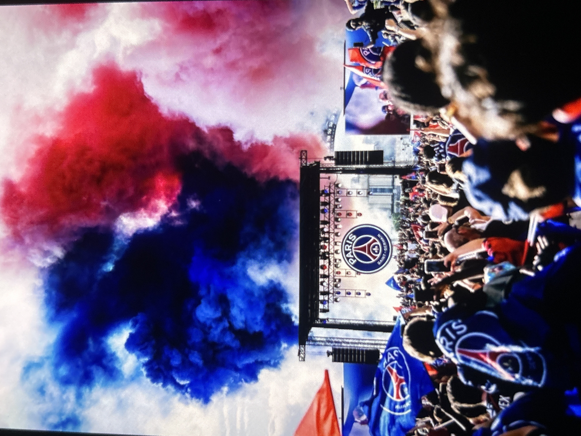 The image captures a dynamic outdoor gathering, likely a celebration related to the Paris Saint-Germain (PSG) football club. A large crowd of people occupies the right side of the frame, many of whom are seen wearing or holding blue flags and apparel featuring the PSG logo. In the central area, a prominent stage setup is visible, complete with lighting rigs and a large display screen. The screen clearly shows the official Paris Saint-Germain crest, which includes the club's name, the Eiffel Tower, and a fleur-de-lis symbol.
To the left of the stage and crowd, dense plumes of red and blue smoke billow upwards, filling a significant portion of the sky. This vibrant smoke, likely from flares or pyrotechnics, adds a dramatic and celebratory atmosphere to the scene. The event appears to be taking place during the daytime, with parts of the sky visible behind the smoke and structures. While individual faces in the crowd are mostly indistinct, the overall impression is one of excitement and collective enthusiasm. Some red flags are also discernible among the blue and white flags held by the attendees.