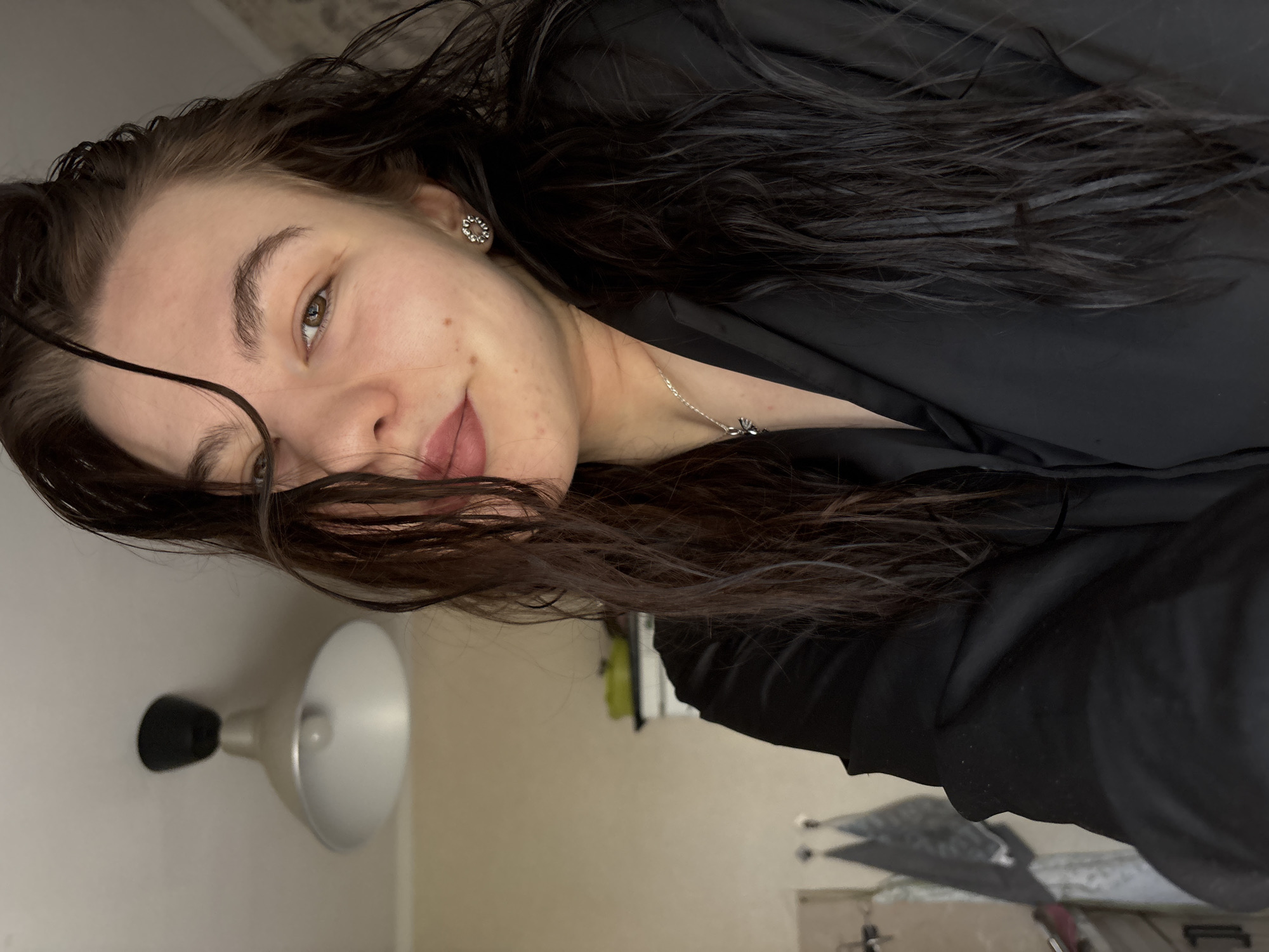 A selfie image shows a woman with long, dark hair, brown eyes, and fair skin. She is wearing a black top and a silver necklace. An earring is visible in her left ear. The background is a room with a lamp mounted on the wall. The location is presumed to be in Perm, Russia, but there is no information in the image to confirm that specific location.