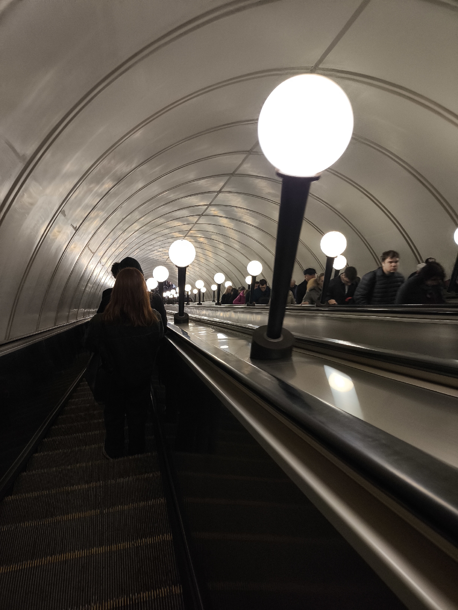 The image shows the interior of a Moscow metro station, likely one of the older, deeper stations known for their architectural features. People are riding up a long escalator. The walls and ceiling are arched and lined with metal or similar material, reflecting light from globe-shaped lamps lining the escalator. People are dressed in casual winter or fall attire. The escalator steps are textured. The scene suggests a typical commute in Moscow, with people traveling between levels of the metro system.