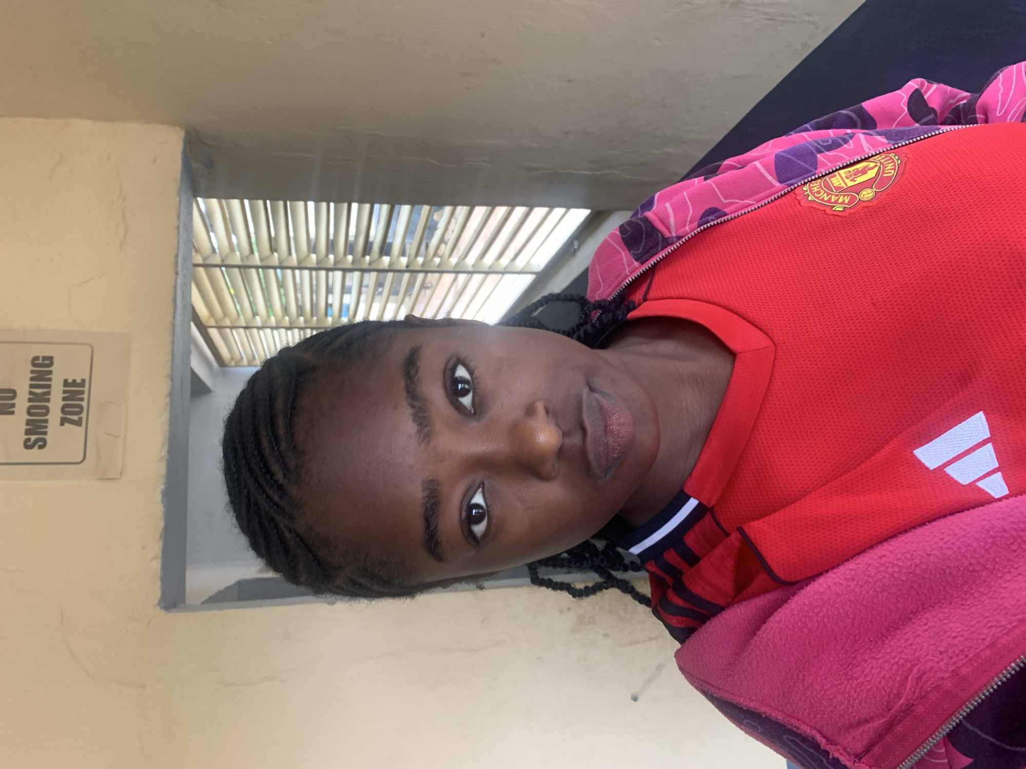A young woman with dark skin and braided hair is captured in a medium close-up, portrait-style photograph. She looks directly into the camera with a neutral expression. She is wearing a red short-sleeved sports jersey, which features a yellow Manchester United crest on the left chest and a white Adidas logo on the right side. Over this, she has a vibrant pink, purple, and black patterned zippered jacket with a visible bright pink fleece lining.
The background suggests a semi-outdoor or sheltered concrete structure. To her left, there's a light-colored wall with a white sign that reads "NO SMOKING ZONE" in black, vertically oriented text. Behind her head and extending to the right, a slatted or louvered structure, possibly a roof or awning, allows daylight to filter through, indicating it is daytime. The architectural elements and the "NO SMOKING ZONE" sign point to a public or facility setting, consistent with the provided context of "New Stadium, Nigeria." The image is rotated 90 degrees counter-clockwise.