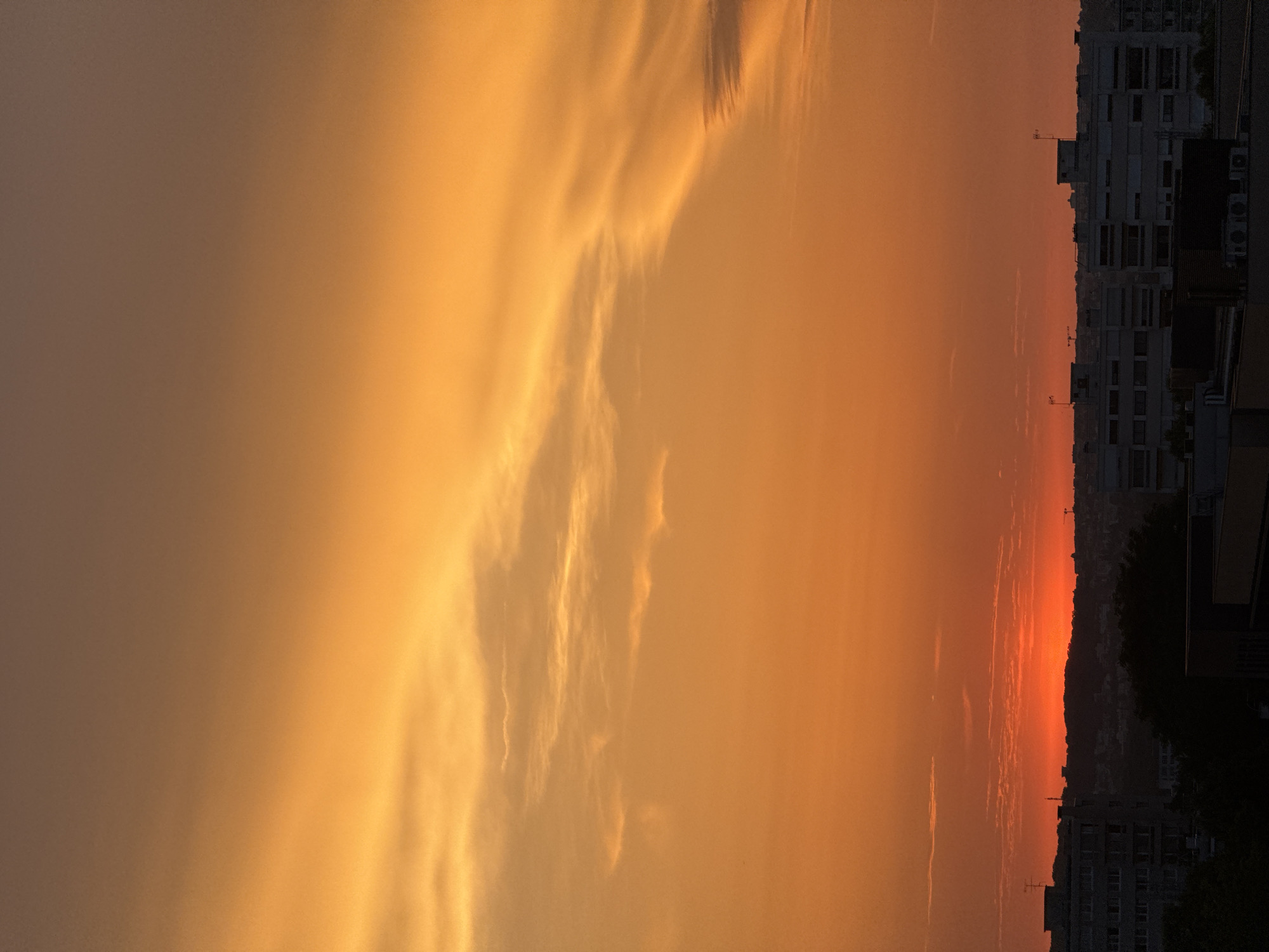 This is a vertical, medium-wide shot of a sunset over a cityscape. The sky dominates the upper two-thirds of the frame, transitioning from a pale brown at the top to a vibrant orange and red near the horizon. Wispy, elongated clouds are visible in the upper sky, painted with shades of orange. The lower third of the image features a silhouetted cityscape. Buildings, some appearing to be apartment blocks with visible windows, line the horizon. A dark, undulating line of hills or a distant elevated terrain is visible behind the buildings, with the brightest red and orange hues of the sunset concentrated near its crest. The foreground buildings are dark silhouettes, obscuring any details of the ground level. The overall impression is of an urban environment at dusk, bathed in the warm, dramatic light of the setting sun.
