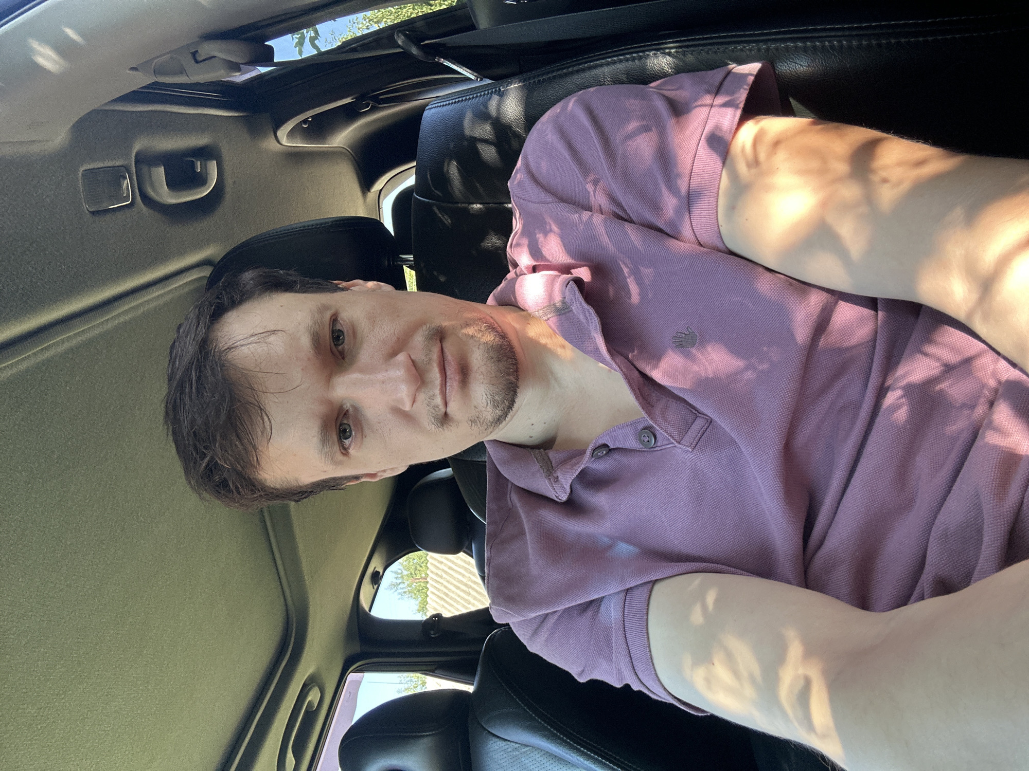 A man is seated in the driver's seat of a car, with the camera positioned in front of him. He has short brown hair and is wearing a purple polo shirt. The car's interior is visible, including the dashboard, sunroof, and passenger seats. Sunlight streams into the car, casting shadows on the man's face and clothing. Outside the car, a fence and trees are visible under a clear blue sky, indicating a rural or suburban setting. The image was taken in Shatava, Ukraine.