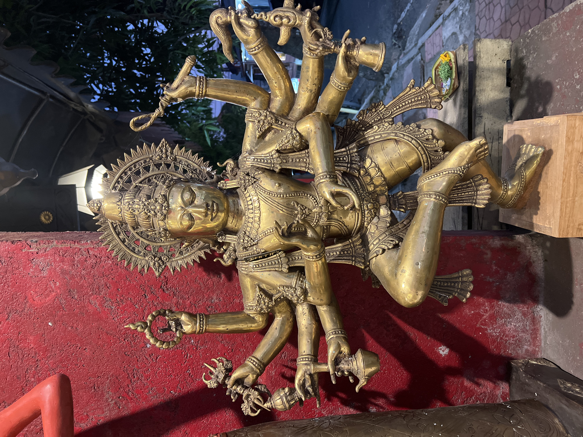 An outdoor photograph, likely taken at dusk or night, depicts a prominent golden, multi-armed statue of a deity against a red textured wall and an outdoor street scene.
The central subject is a highly detailed, polished golden statue, approximately 1.5 to 2 meters tall, depicting a Hindu or Buddhist deity in a dynamic, dancing pose. The deity features a serene facial expression with closed eyes, crowned by an elaborate, radiating halo. The statue possesses eighteen arms: two main arms are positioned at the chest in a mudra, while eight additional arms extend from each side of the torso. Each of the sixteen extended hands holds a distinct object: on the left side (from top to bottom), a noose/lasso, a small bell, an ornate pitcher, a trident-like object, and another ornate pitcher; on the right side (from top to bottom), a stylized club or conch, a coiled rope/whip, a floral or flame-like emblem, a small bell, and a larger bell. The deity's attire includes intricate jewelry, armlets, anklets, and a decorated lower garment. The statue is mounted on a light wooden block, which rests on a darker, possibly concrete, base.
The background consists of two distinct areas. To the left, a vibrant, rough-textured red wall displays shadows cast by the statue's arms and head, along with a partial view of a smooth, red, curved object (likely part of another statue) in the upper left corner. To the right, an outdoor environment is visible under a dark sky, featuring dark green foliage from trees and artificial light sources, including a white rectangular lantern and distant streetlights. A dark, corrugated roof section overhangs from the top right. On the ground to the right, a paved sidewalk with red interlocking bricks is visible. A small, traditional Balinese *canang sari* offering, consisting of woven palm leaves with colorful flowers and leaves, is placed on a low wooden platform beside the statue's base. The lighting suggests artificial illumination in a low-light setting. The statue type, offering, and architectural elements are consistent with the cultural landscape of the Ubud District, Indonesia.