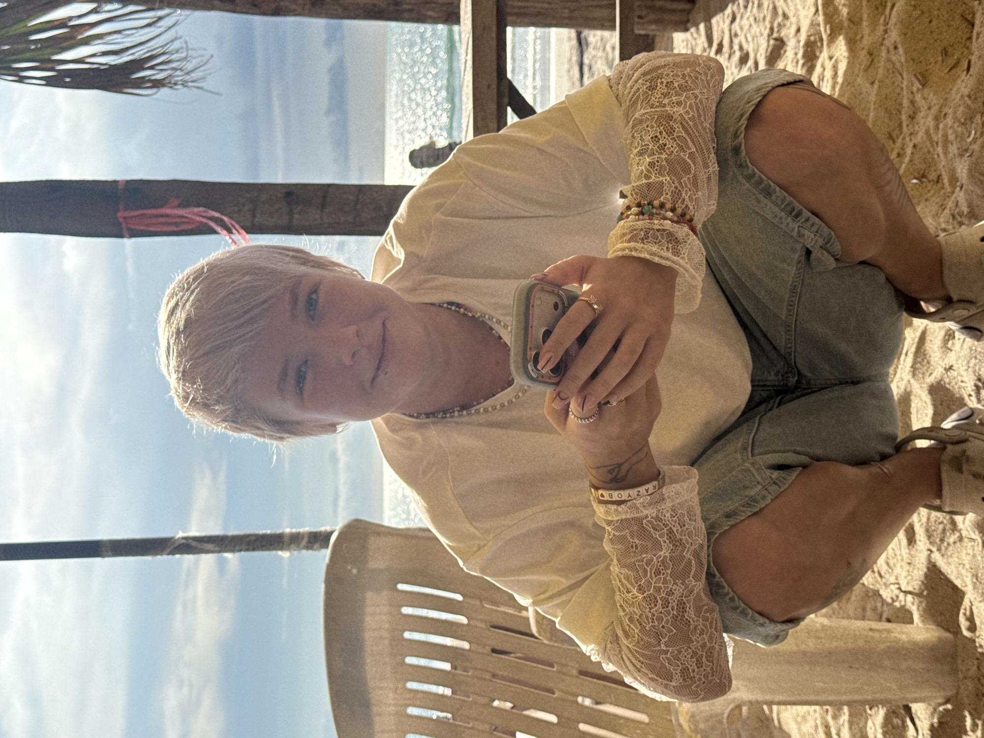 A young person with short, light-colored hair is crouching on a sandy beach, holding a smartphone in their hands. They are wearing a loose, light-colored t-shirt with white lace sleeves, denim shorts, and light-colored shoes. The person has several bracelets on their left wrist and rings on their fingers. Behind them, a beach chair and wooden structures are visible, along with palm trees and the ocean under a cloudy sky. The setting appears to be a beach resort in Choeng Thale, Thailand, indicated by the presence of palm trees, the ocean, and typical beach amenities. The overall mood is relaxed and casual, with the person seemingly engaged with their phone.