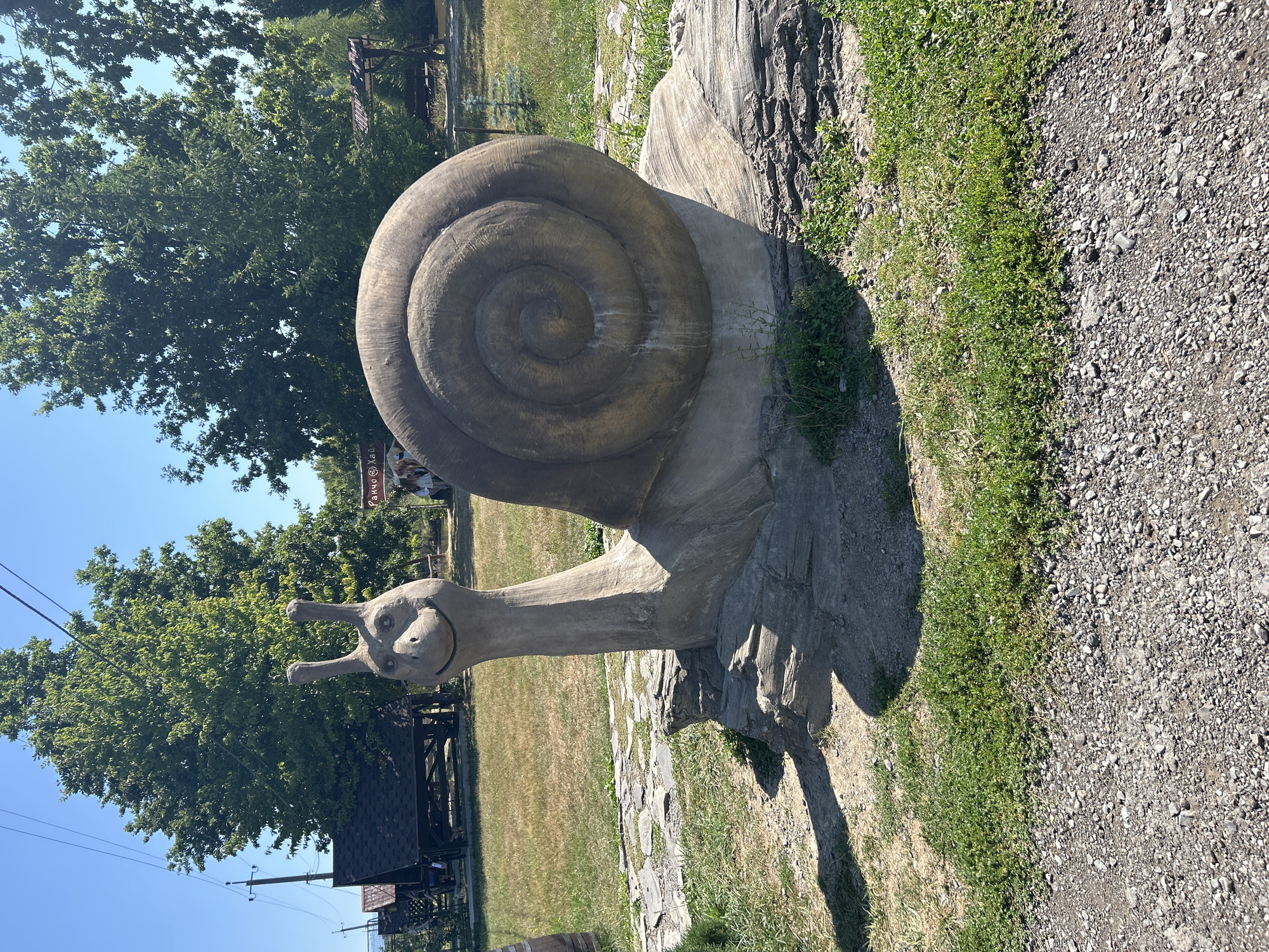 The image captures a concrete sculpture of a snail in an outdoor setting. The snail sculpture is positioned on a base made of grey stones and what appears to be a large log. It has a distinct spiral shell and a head with two antennae, featuring a smiling expression. The sculpture is situated on a grassy area, with a gravel path visible in the foreground. In the background, there are trees, a gazebo-like structure, and a sign with Cyrillic text. The sky is clear and blue, indicating a sunny day. The environment suggests a park or recreational area, likely in Lypcha, Ukraine, given the visual context and typical rural landscaping. There are a few indistinct figures in the background, near the sign.