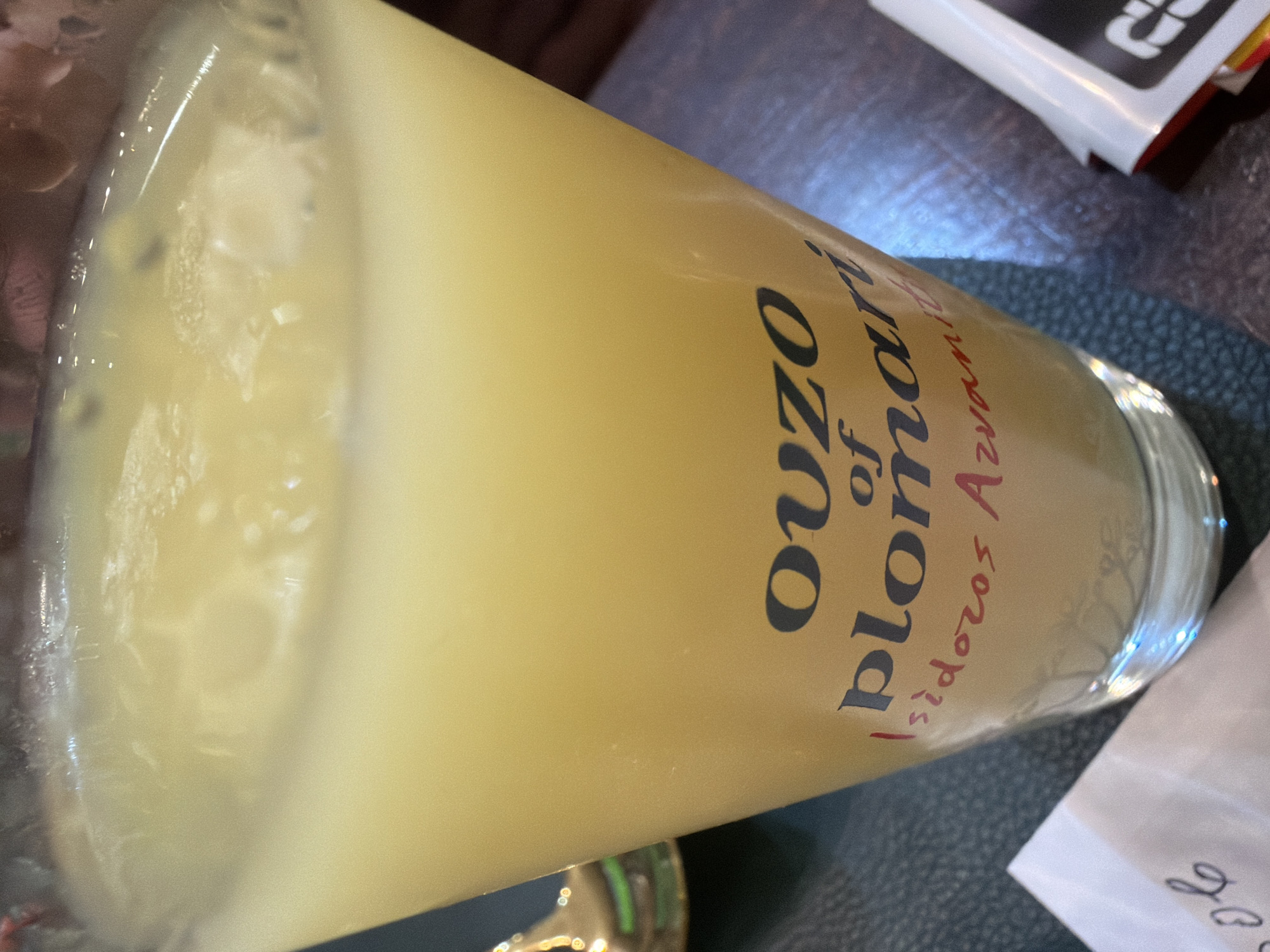 A close-up, eye-level, interior shot shows a glass of pale yellow liquid with ice cubes and foam. The glass is cylindrical and tapers slightly towards the top. Dark lettering on the glass reads "OUZO of plomari Isidóros Arvanítis". The glass sits on a dark, textured surface, possibly a table. To the left, a white paper with handwritten dark cursive text is partially visible. To the right, a white object with red and black accents is also partially visible. The background is dark and out of focus, suggesting an indoor setting.