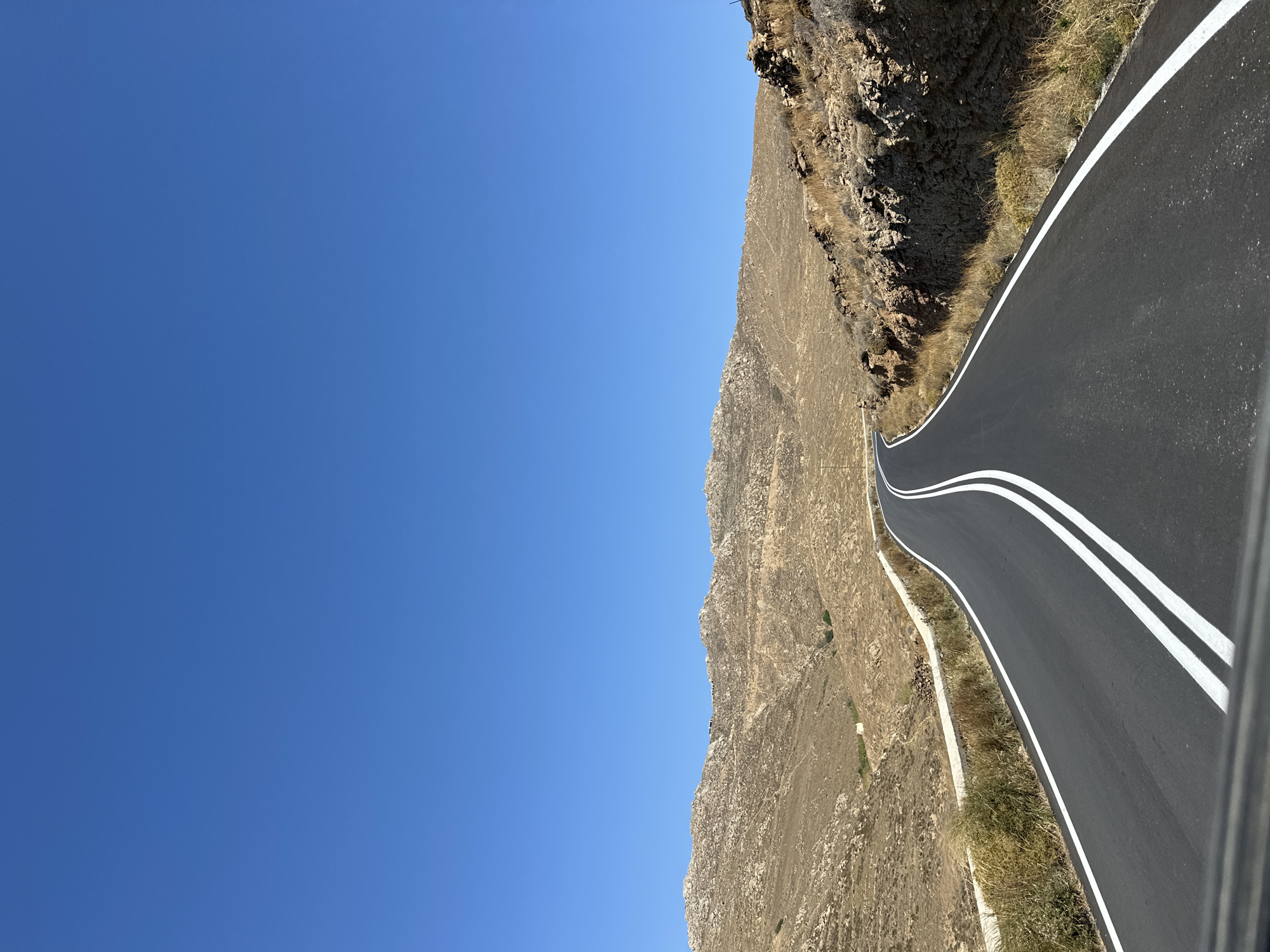 The image shows a winding asphalt road with double white lines in the center, disappearing over a hill. The road is flanked by dry, rocky terrain with sparse vegetation. In the background, there are rolling hills and mountains under a clear blue sky. The scene appears to be a rural area in Mykonos, Greece, likely on a sunny day. There are no people or vehicles visible in the frame.