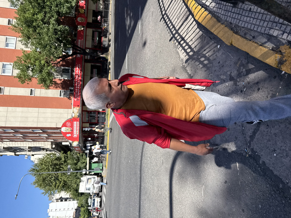 A man with gray hair, wearing a tan shirt and red jacket, stands on a street. He's in the middle of the road, near a curb with yellow paint. Buildings and trees are visible in the background, along with parked vehicles. The scene is bathed in sunlight.