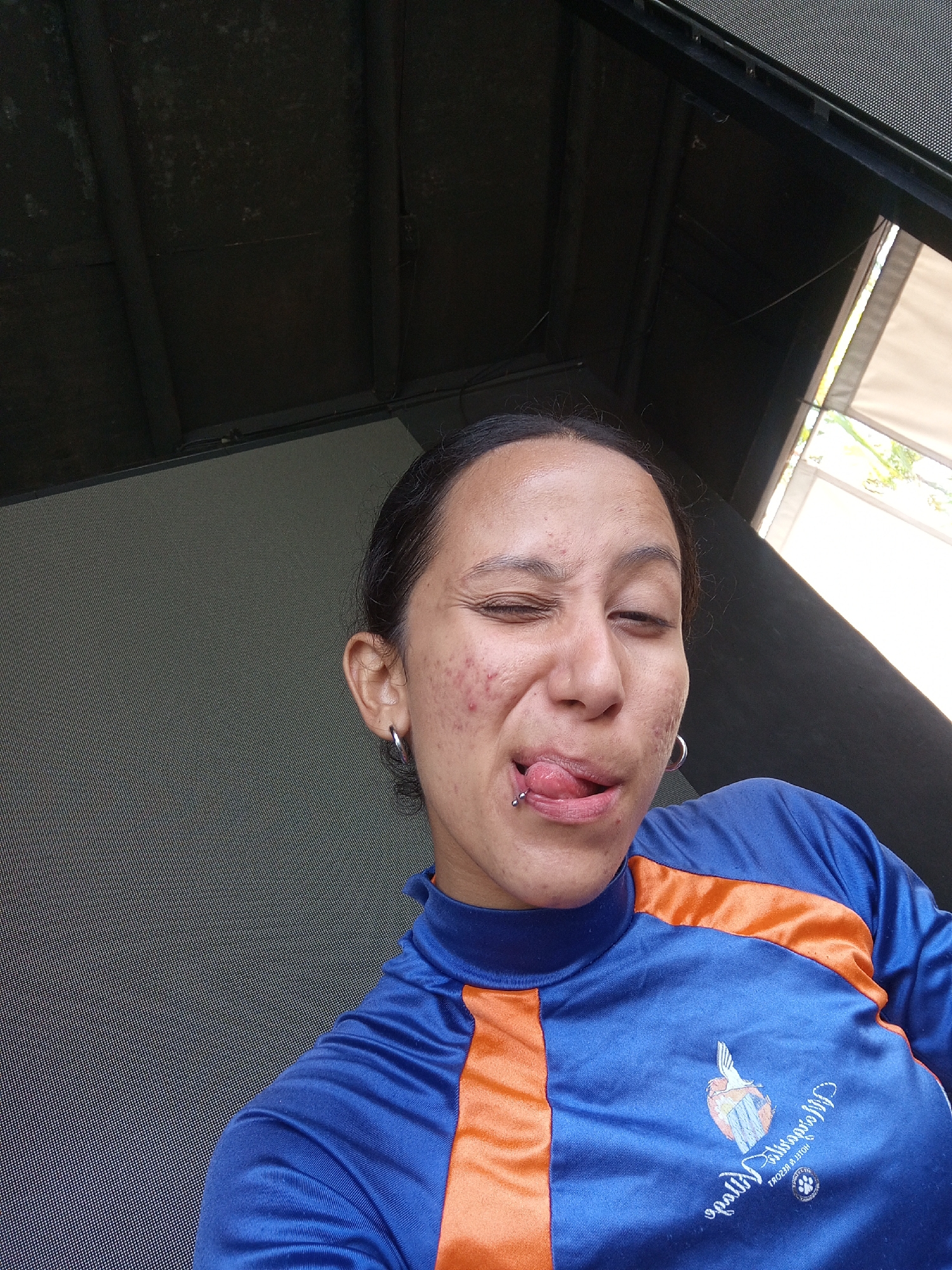 A light-skinned person with dark hair pulled back is centered in the frame, sticking their tongue out and winking. They have a silver barbell piercing in their tongue and silver hoop earrings. Their face has visible blemishes. They are wearing a blue and orange athletic shirt with a logo on the front. The logo appears to read "Hesperia Isla Margarita Hotel & Resort". Based on the hotel name, the location is likely Porlamar, Venezuela. The background shows a partially visible window and an indoor ceiling. The room appears to be enclosed, possibly a covered patio or indoor area.