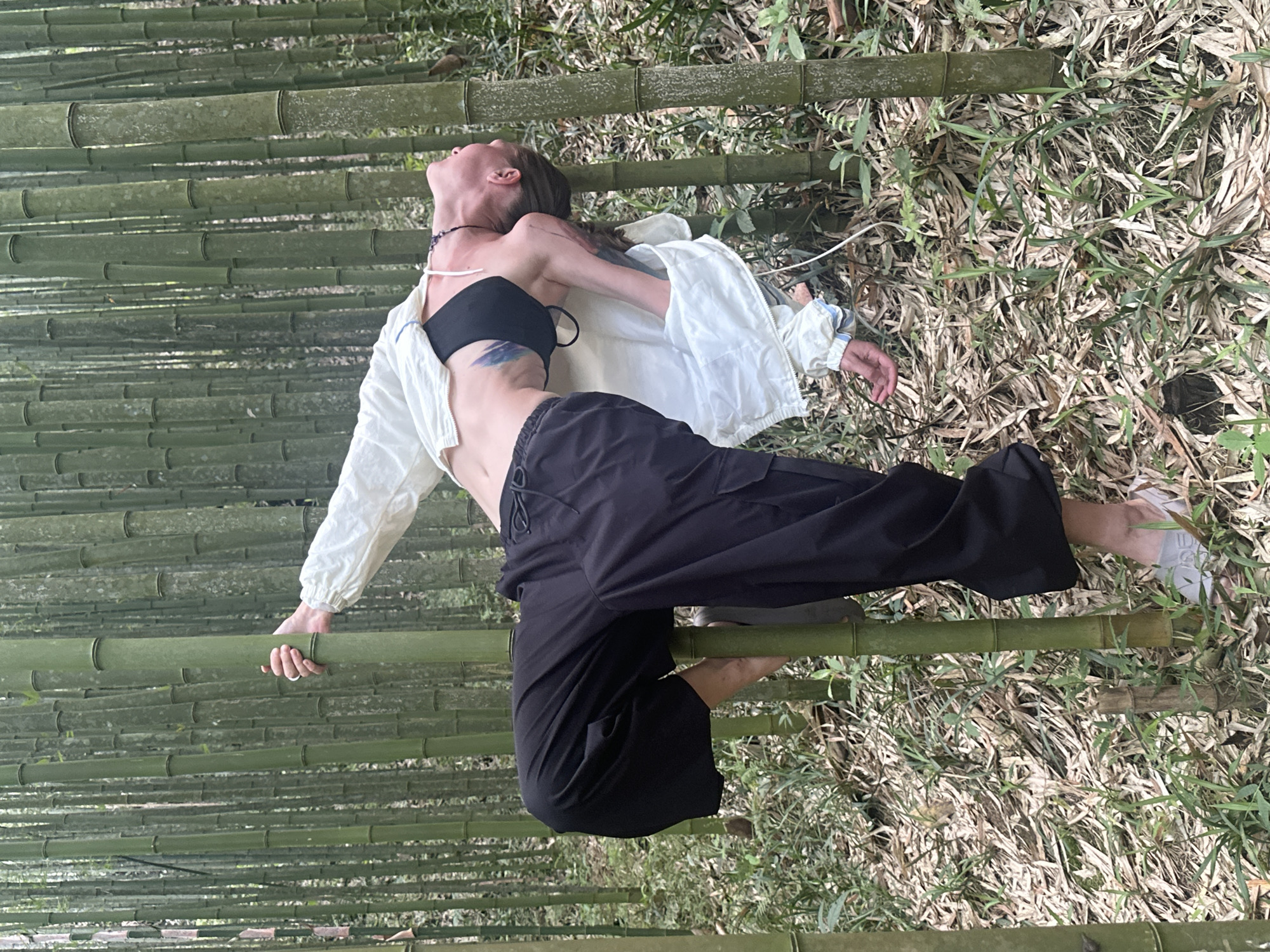 The image shows a woman in a black strapless top, a white jacket, and black pants posing amongst bamboo stalks and dried vegetation. She is grasping a bamboo stalk with her left hand, and her head is tilted back. Her right arm is extended to the side, and she is barefoot. The bamboo stalks are aligned horizontally. The ground is covered in dry leaves and some green foliage. The location is likely a bamboo forest, with natural light illuminating the scene. There is no information to associate the image with Ngã Ba Kim, Vietnam.