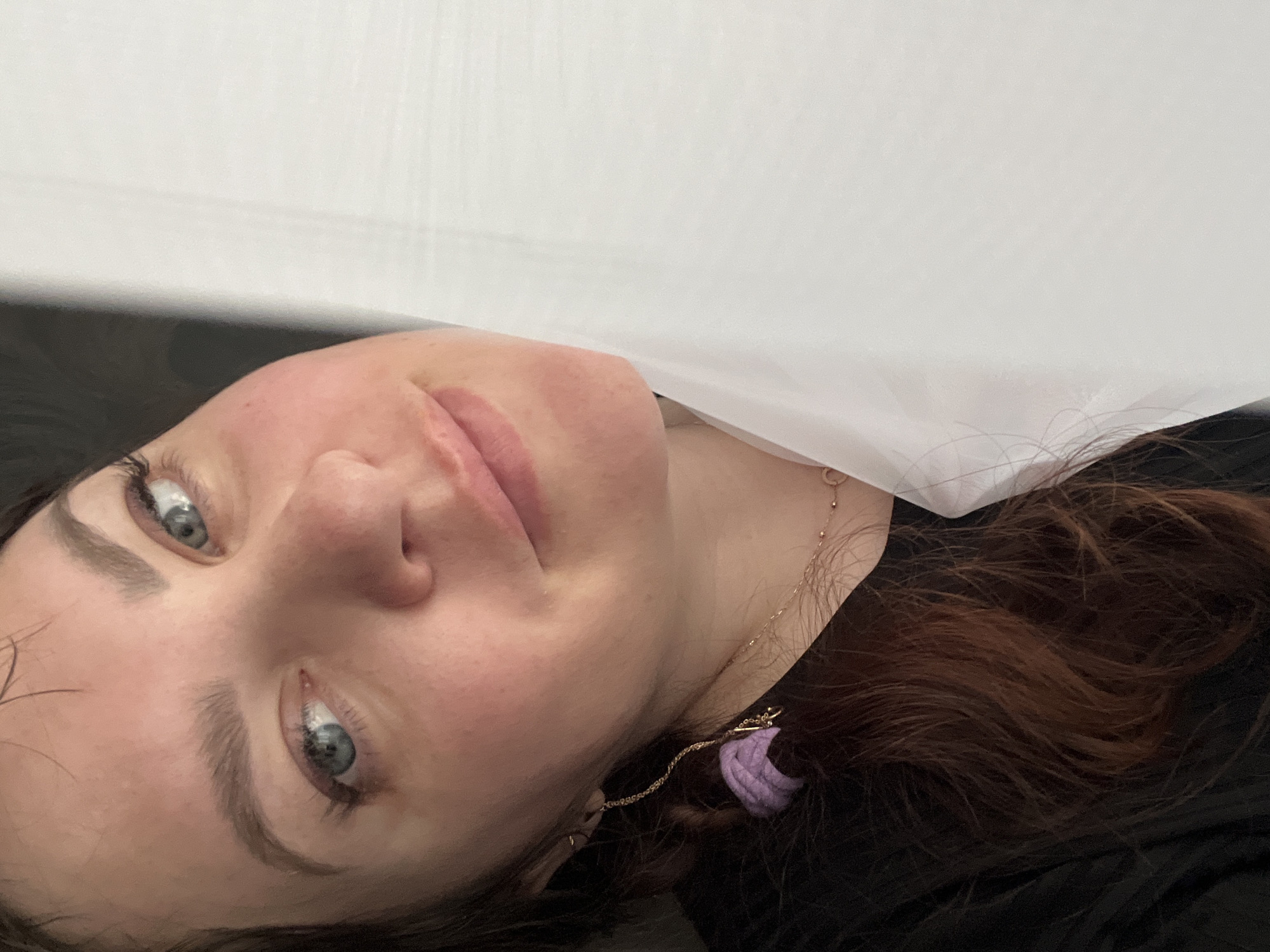 A young woman with blue eyes and dark brown, wavy hair is depicted in a close-up, slightly overhead shot. She has light skin with a natural flush on her cheeks and defined eyebrows. Her lips are closed in a soft, gentle smile, and she is looking directly towards the camera. She appears to be lying down, possibly on her back, with her head rotated.
She is wearing a black top or garment. A delicate gold chain necklace with a small pendant is visible around her neck. On the right side of her head, blending into her hair, is a distinctive purple, textured hair accessory, possibly a scrunchie, attached to another gold chain. A small, simple gold earring is also visible in her left ear (on the right side of the image).
The background is simple, divided between a dark surface on the left and a bright white surface on the right, which could be bedding, a wall, or a ceiling, indicating an indoor setting. There is no visible text. The overall mood conveyed by her expression is calm and content. The location context is Noviy, Russia.