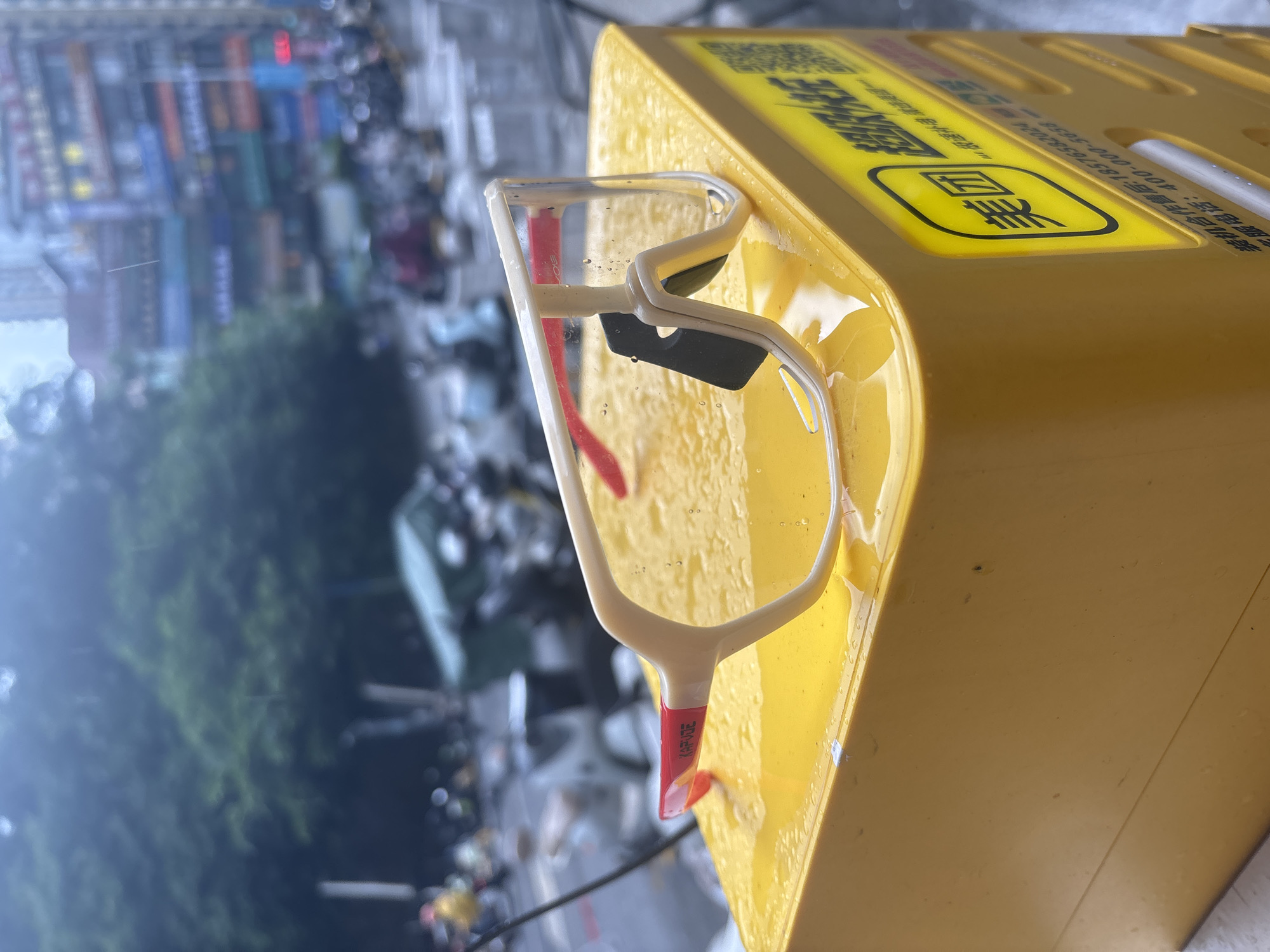 A pair of oversized, wraparound sunglasses with a white frame and clear lenses, accented with red on the inside of the arms, rests on a yellow container. The sunglasses are wet, with visible water droplets on the lenses and frame. The yellow container has a graphic of a black rectangle within a rounded square with Chinese characters above it, and more Chinese text below. The background is blurred, showing a street scene with trees, buildings, and parked motorcycles. The presence of water on the sunglasses and the overall wet appearance of the background suggest it is raining. The Chinese text on the container identifies it as belonging to "Meituan", a prominent Chinese e-commerce and delivery company. The location is specified as Nanhai District, China.