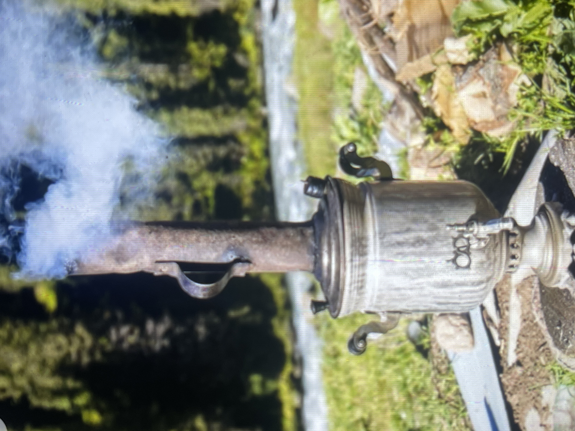 A close-up shot focuses on a rustic, silver samovar situated outdoors. The samovar is a traditional Russian kettle used for heating water and is emitting a plume of white steam that rises from its central chimney. The samovar has ornate handles and a spigot. In the background, out of focus, is a fast-flowing river, and beyond that, a dense forest of evergreen trees. The ground in the foreground is covered with green grass and scattered rocks. The scene suggests a natural, possibly mountainous, environment. The image quality is low, with visible pixelation and color noise, making precise identification of location difficult. However, the overall aesthetic is consistent with outdoor activities in natural settings such as those found near Bishkek, Kyrgyzstan.