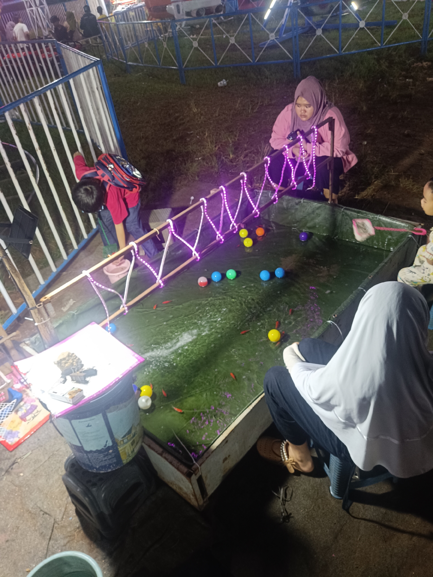 The image depicts an outdoor scene at night, likely at a fair or carnival in Depok, Indonesia. Several people are gathered around a shallow pool of green water, where small red fish are swimming. The pool is illuminated by strings of purple LED lights that run along a wooden walkway spanning the water.
In the foreground, a person wearing a white hijab and dark clothing sits on a chair, facing the pool. To the right, a young child in a patterned outfit is also looking towards the water. In the middle ground, a woman wearing a pink hijab and a pink jacket is crouched down, appearing to observe the fish. To the left, a young boy with a red shirt and a backpack leans over the pool, seemingly engaged in the activity.
Various colored balls are floating on the surface of the water, including yellow, blue, green, and red and white striped ones. A small net is visible near the edge of the pool, suggesting a game or activity where participants might try to catch the fish or balls. In the background, a white fence with blue accents encloses part of the area, and the dim lighting indicates it is nighttime. There is no visible text in the image. The overall atmosphere appears to be one of casual enjoyment and entertainment.